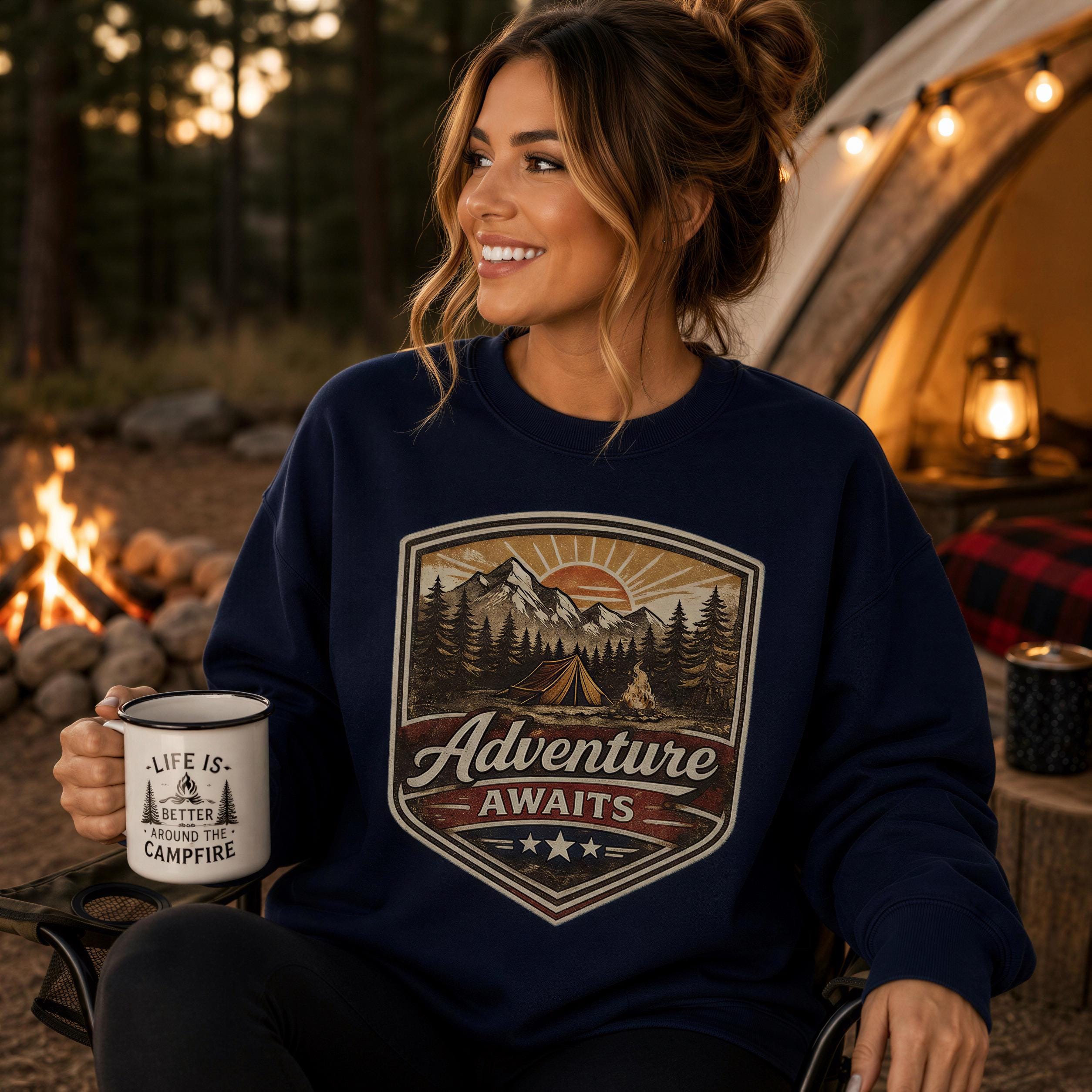 A woman wearing a navy blue sweatshirt with a graphic design of a mountain landscape and camping gear, holding a mug with the text "Life is better around the campfire".