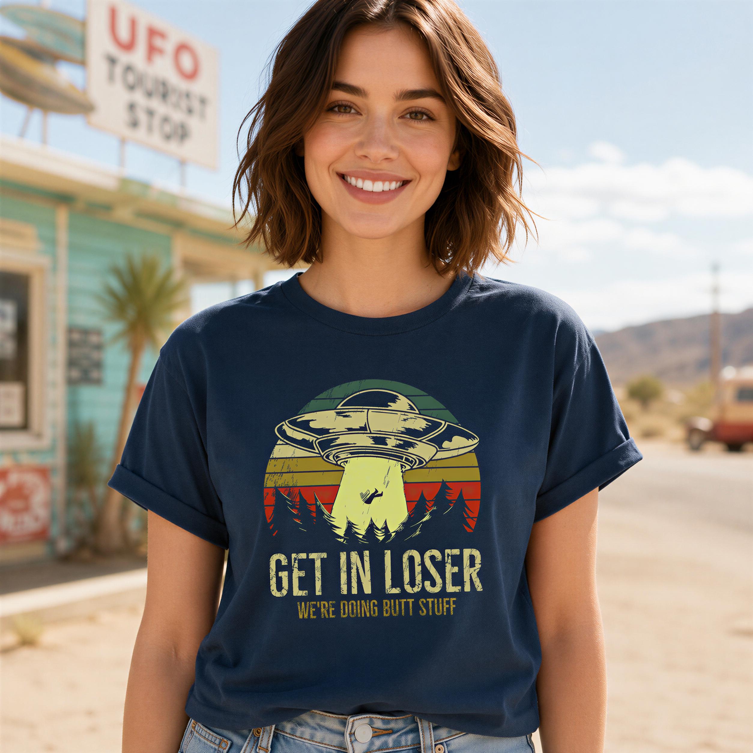 A woman wearing a blue t-shirt with a graphic of an alien spacecraft and the text "GET IN LOSER WE'RE DOING DUTT STUFF" is smiling at the camera.