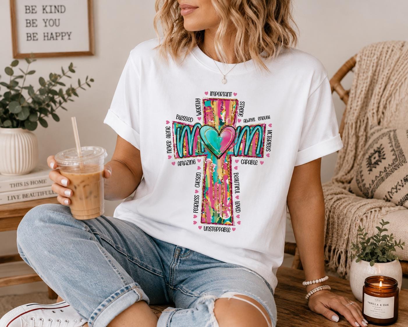 A woman wearing a white t-shirt with a colorful graphic design and holding a drink is sitting on a wooden bench in a room with various decor items.
