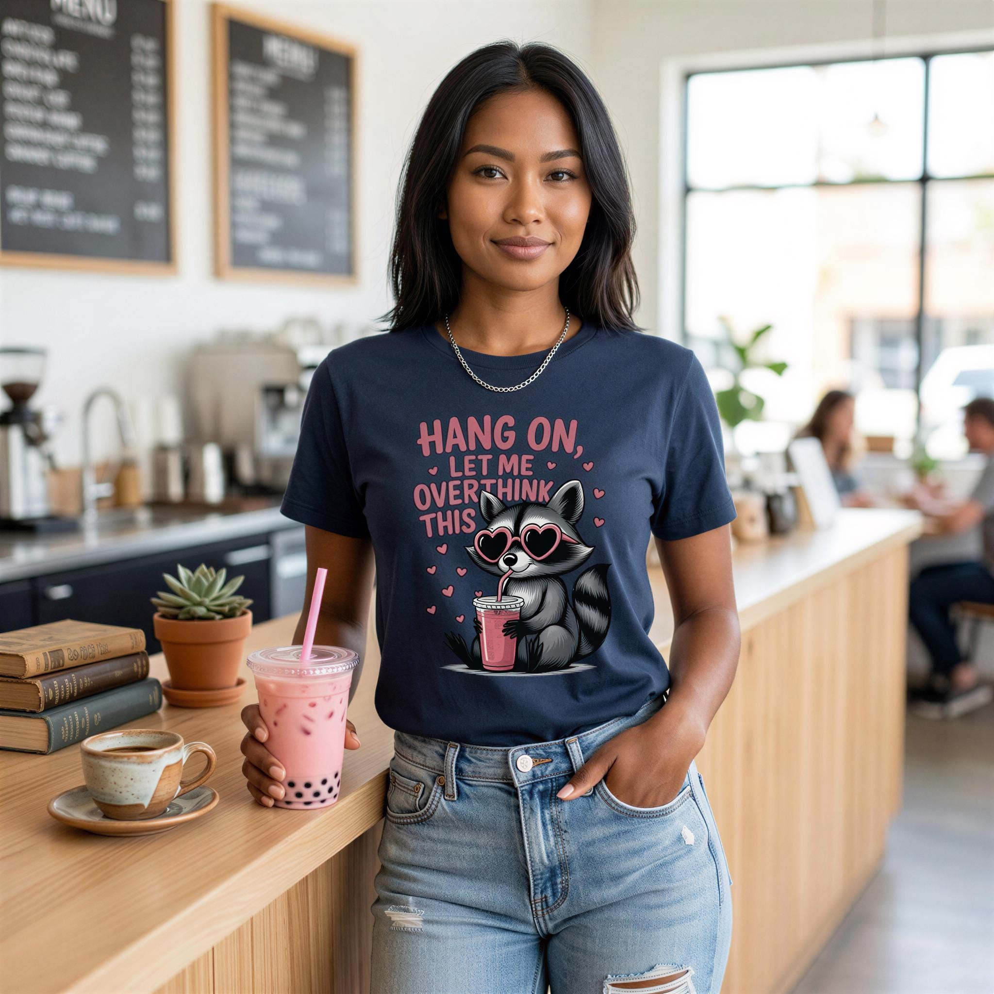 A woman wearing a blue t-shirt with a graphic of a raccoon and the text "HANG ON, LET ME OVERTHINK THIS" is standing in a coffee shop, holding a pink drink with a straw.