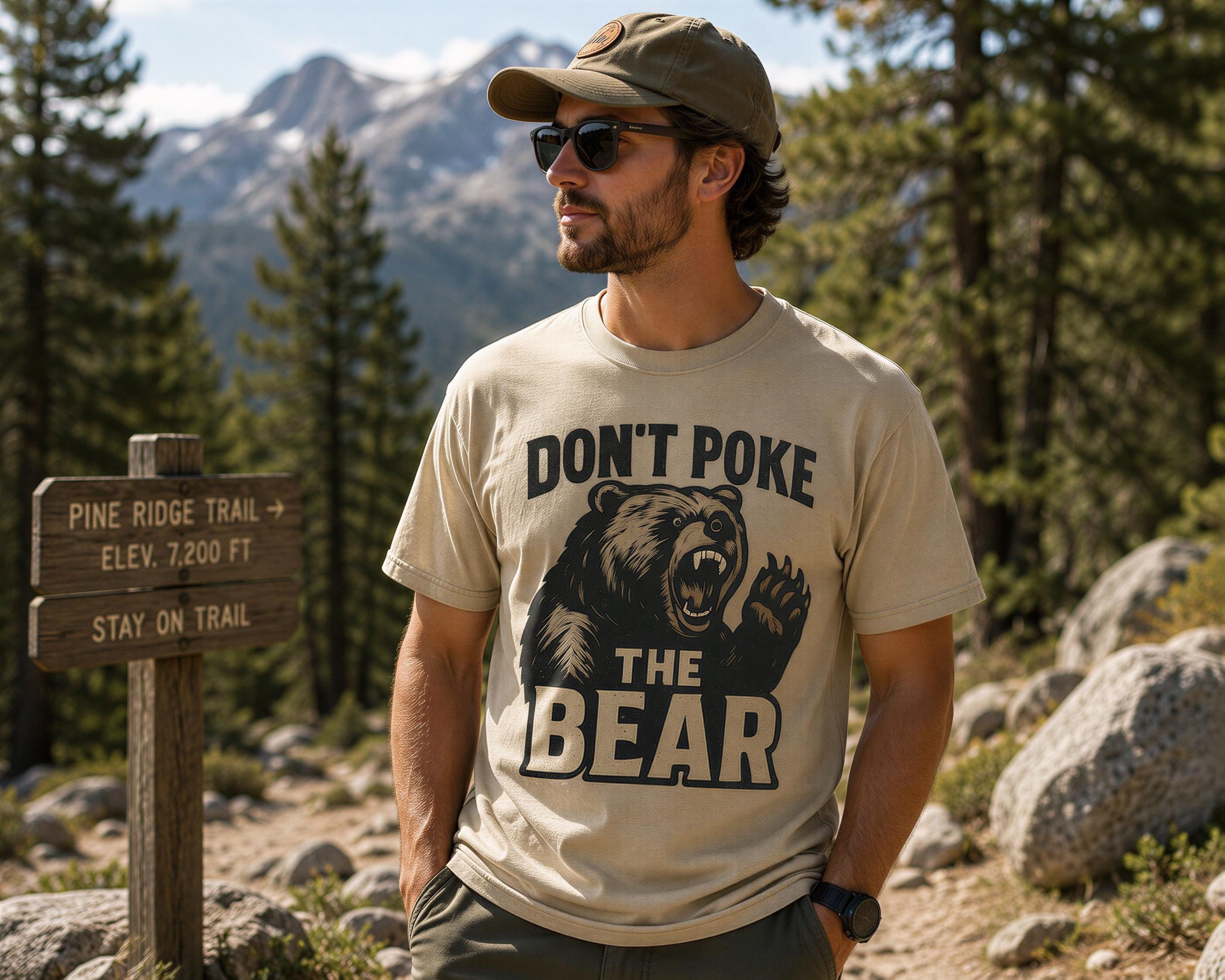 A man wearing a t-shirt with a bear graphic and the text "DON'T POKE THE BEAR" is standing on a trail in a forested area with mountains in the background.
