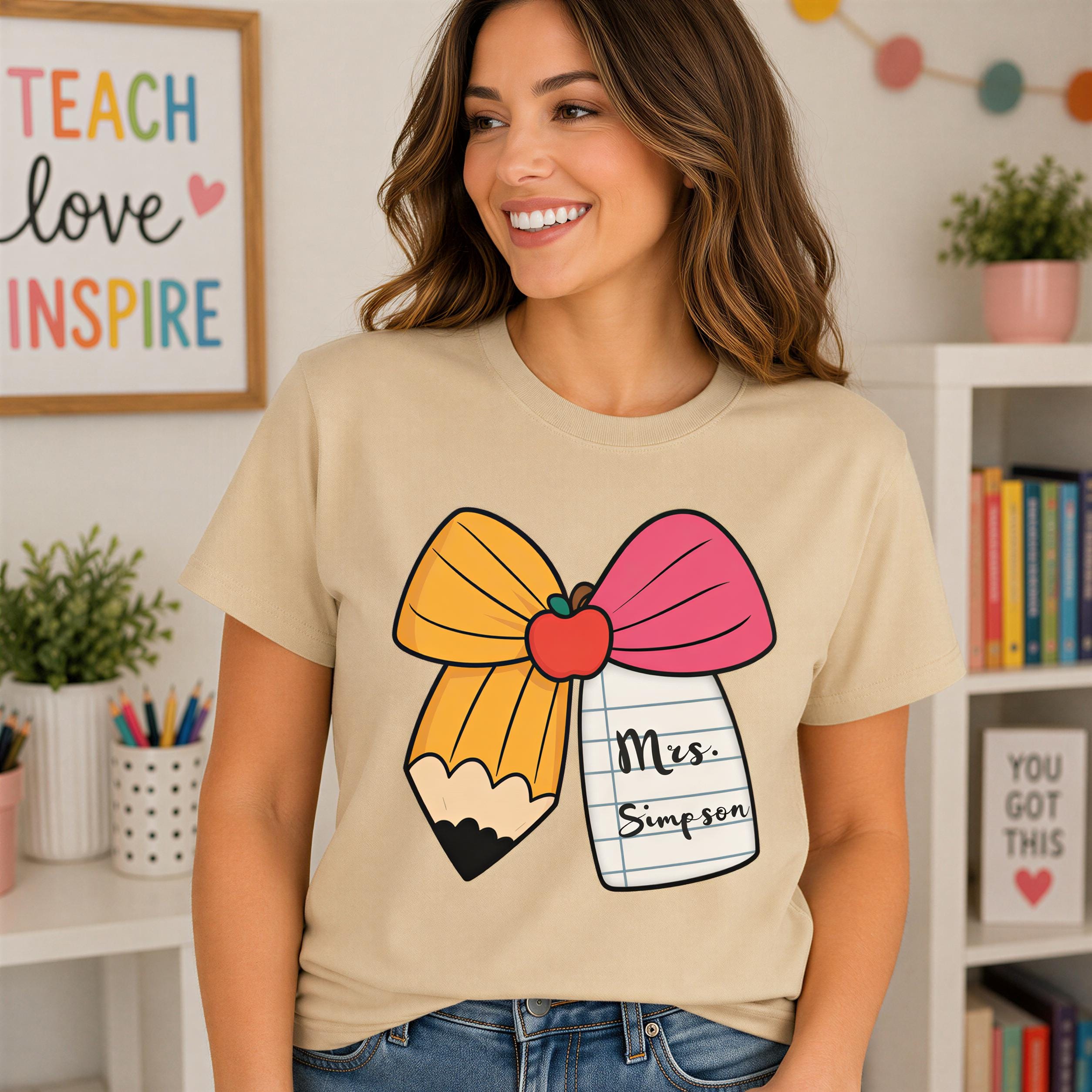 A woman wearing a t-shirt with a pencil and apple graphic, along with the text "Mrs. Simpson" printed on it, smiling in a room with bookshelves and a colorful wall decoration.