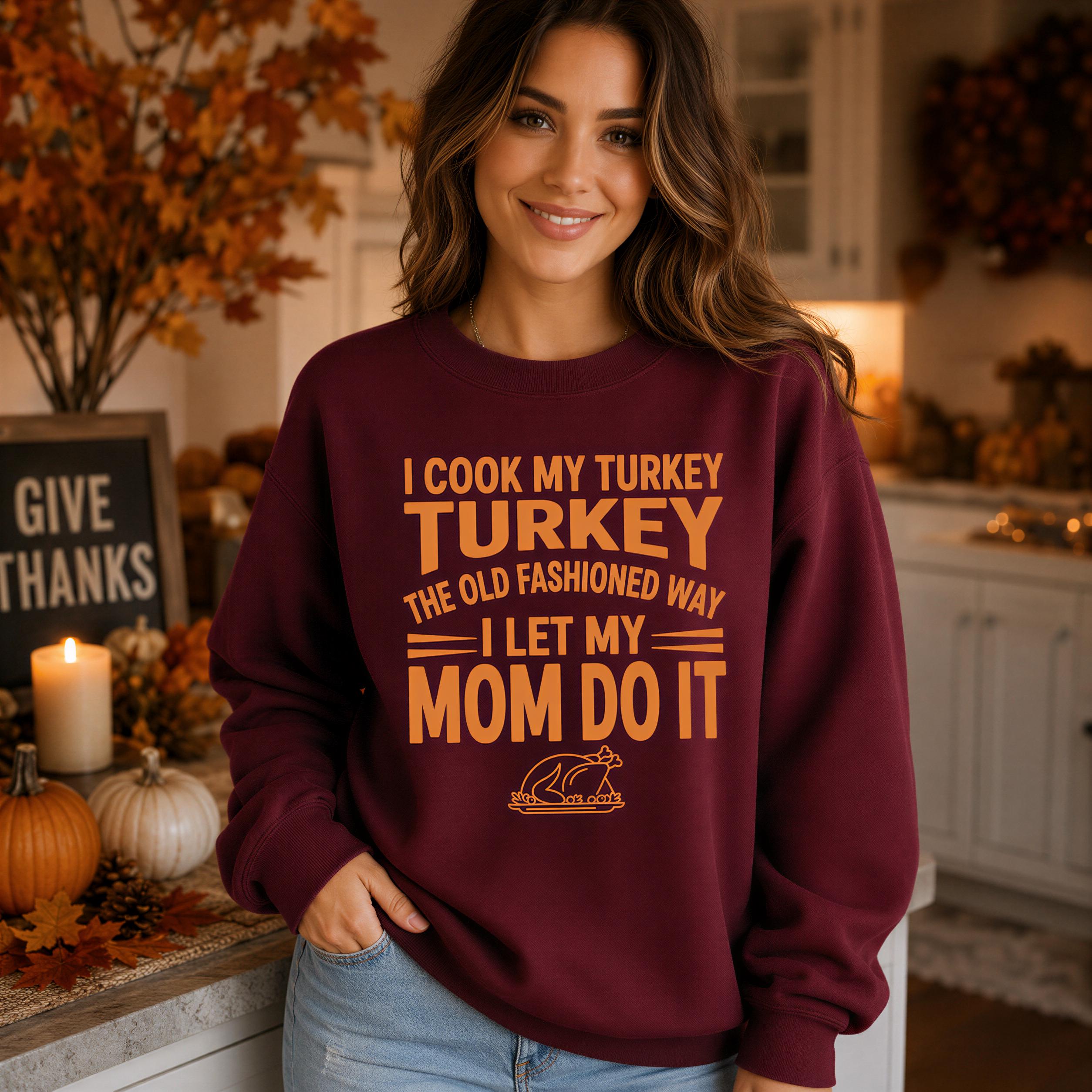 A woman wearing a burgundy sweatshirt with text that says "I cook my turkey the old fashioned way I let my mom do it" standing in a kitchen with fall decorations and a lit candle.