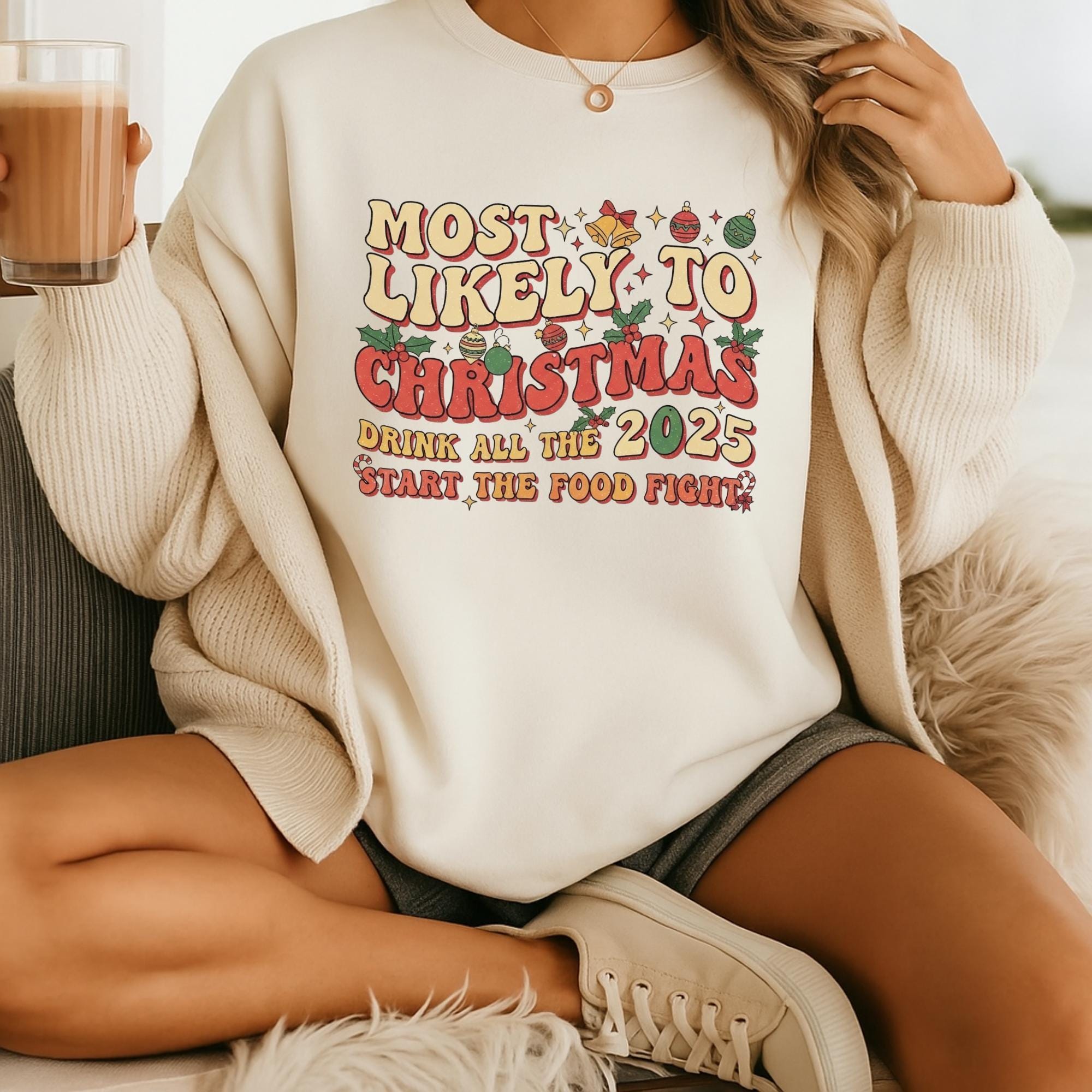 A person wearing a white sweater with a humorous Christmas-themed graphic and text, sitting on a couch and holding a drink.