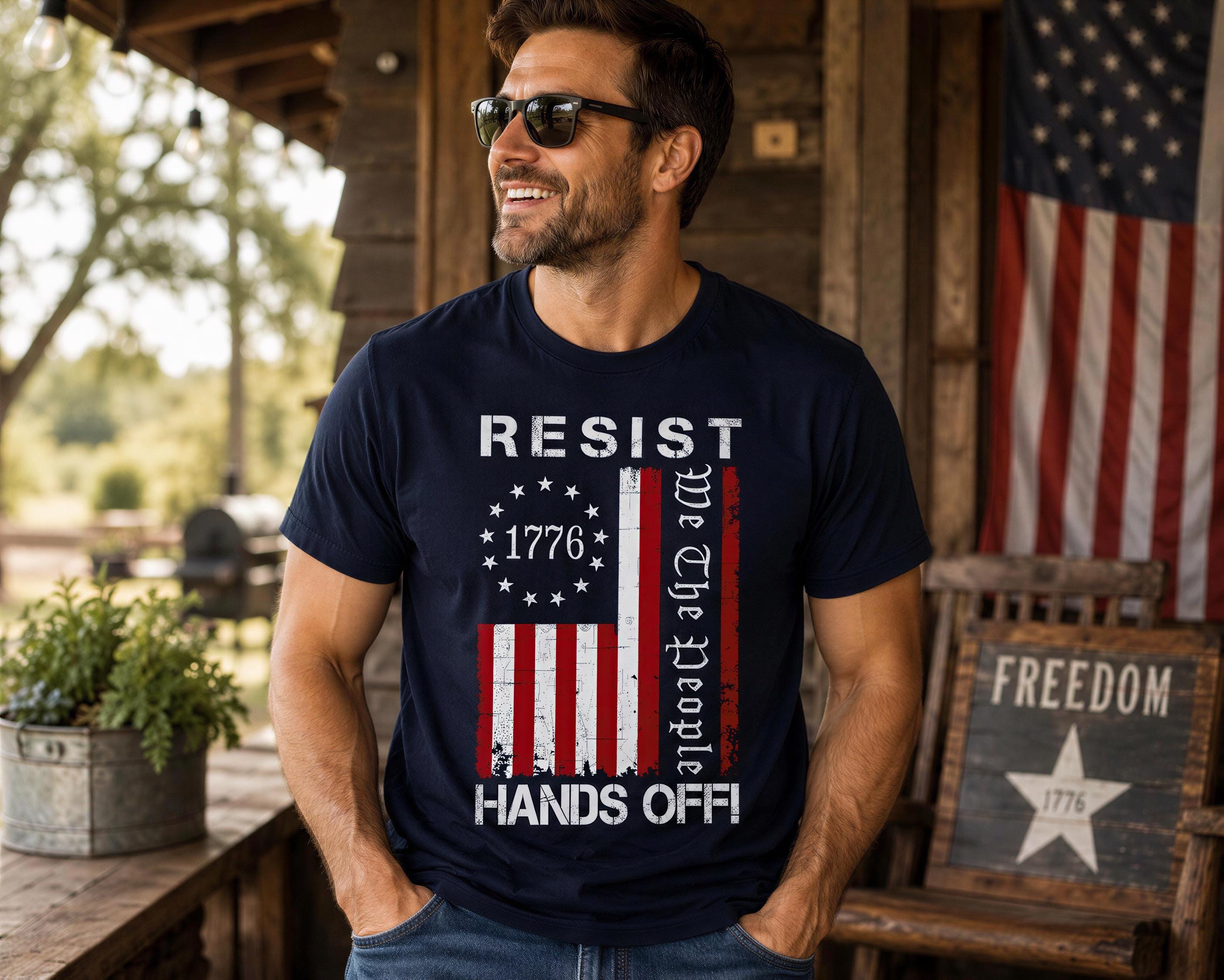 A man wearing a navy blue t-shirt with a patriotic design stands in front of an American flag and a wooden structure, smiling and looking to the side.