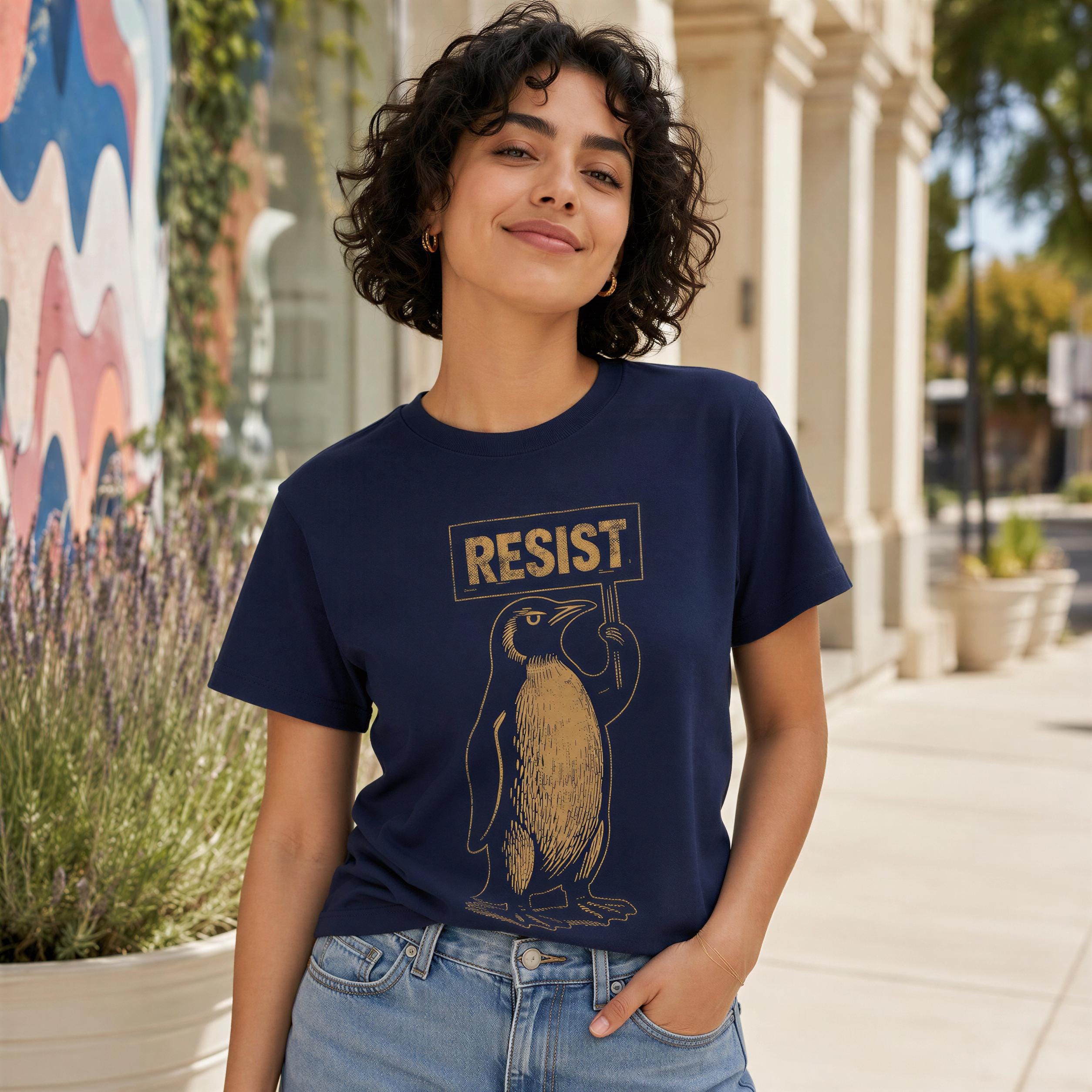 A young woman wearing a navy blue t-shirt with a graphic of a penguin holding a sign that says "RESIST" is standing in front of a colorful mural.