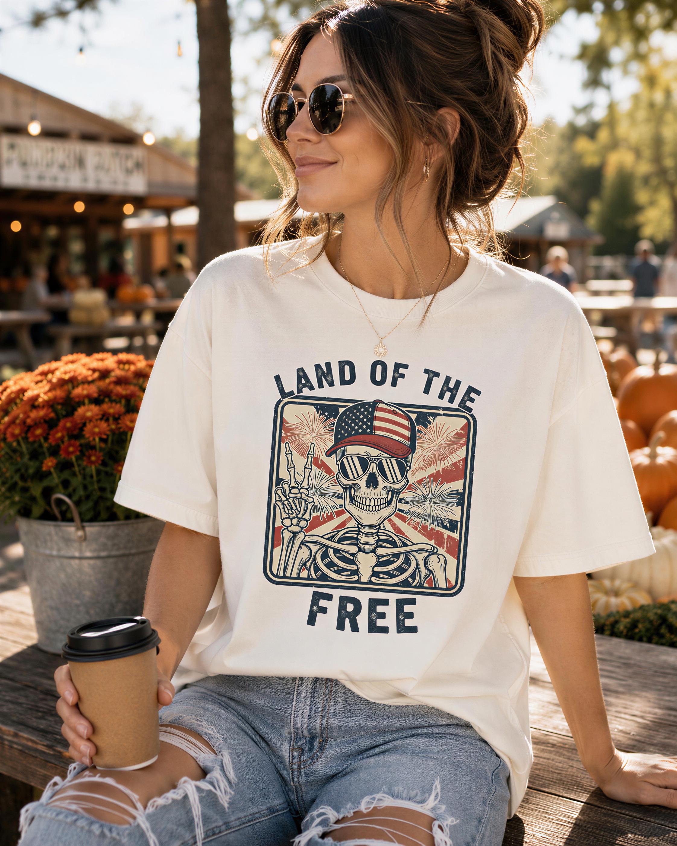A woman wearing a white t-shirt with a graphic design and the text "LAND OF THE FREE" is sitting on a wooden bench, holding a coffee cup.