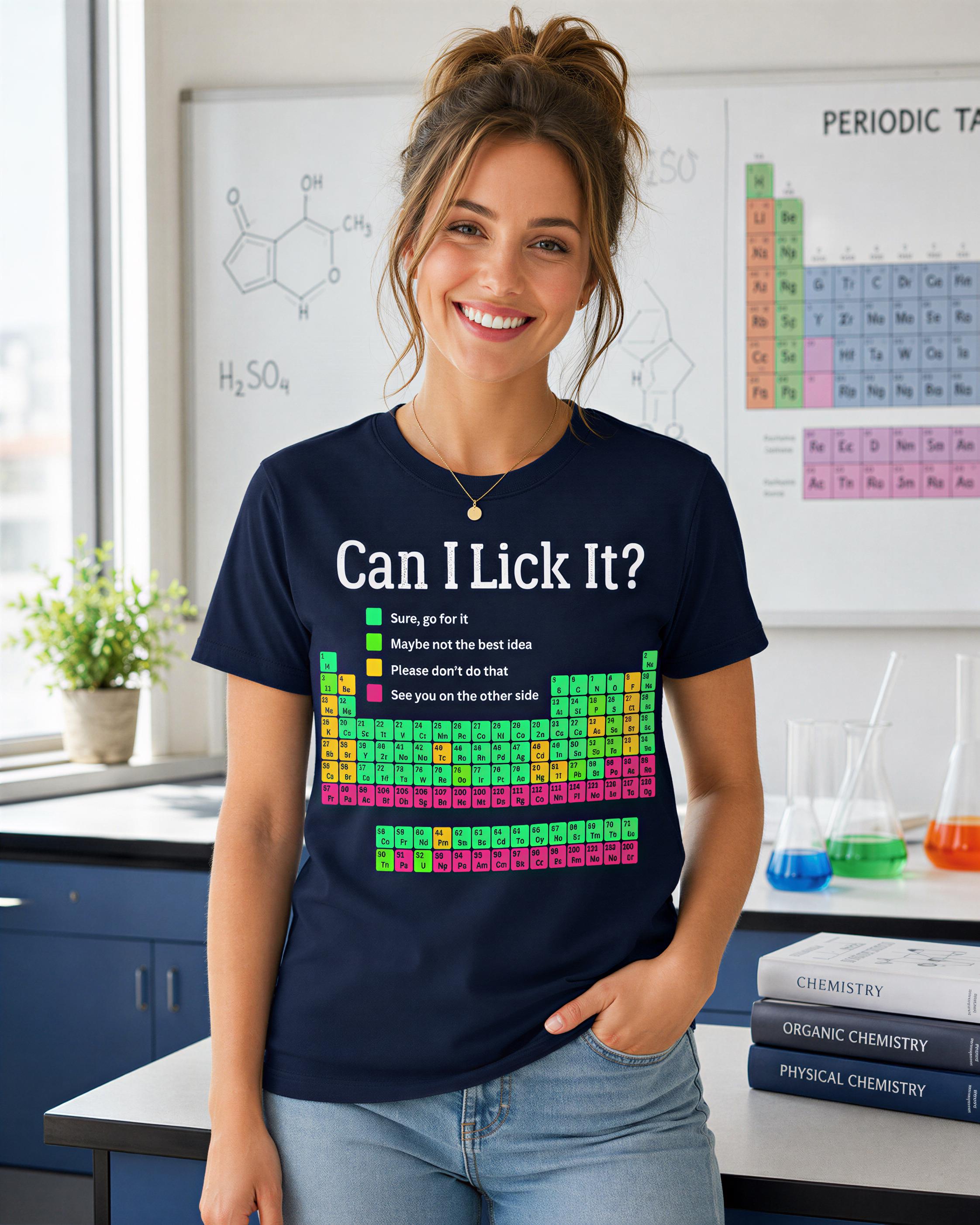 A woman wearing a black t-shirt with a humorous text and a periodic table graphic, standing in a laboratory setting with various scientific equipment and books visible in the background.