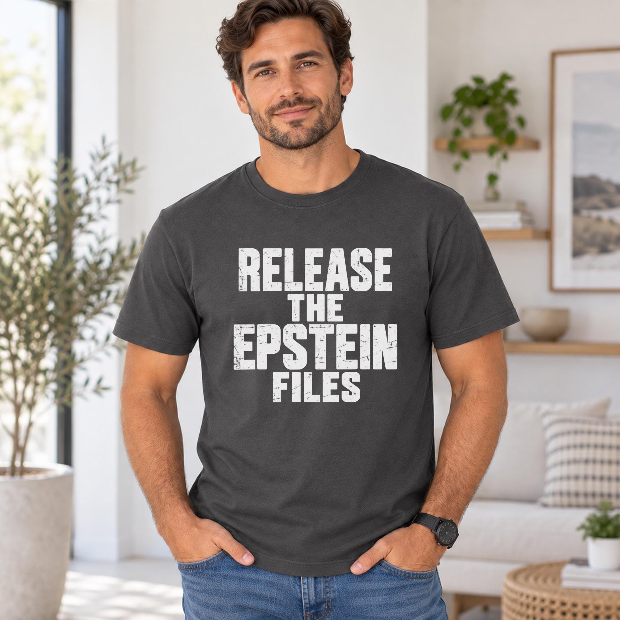 A man wearing a black t-shirt with the text "RELEASE THE EPSTEIN FILES" printed on it, standing in a room with a potted plant and a framed picture visible in the background.