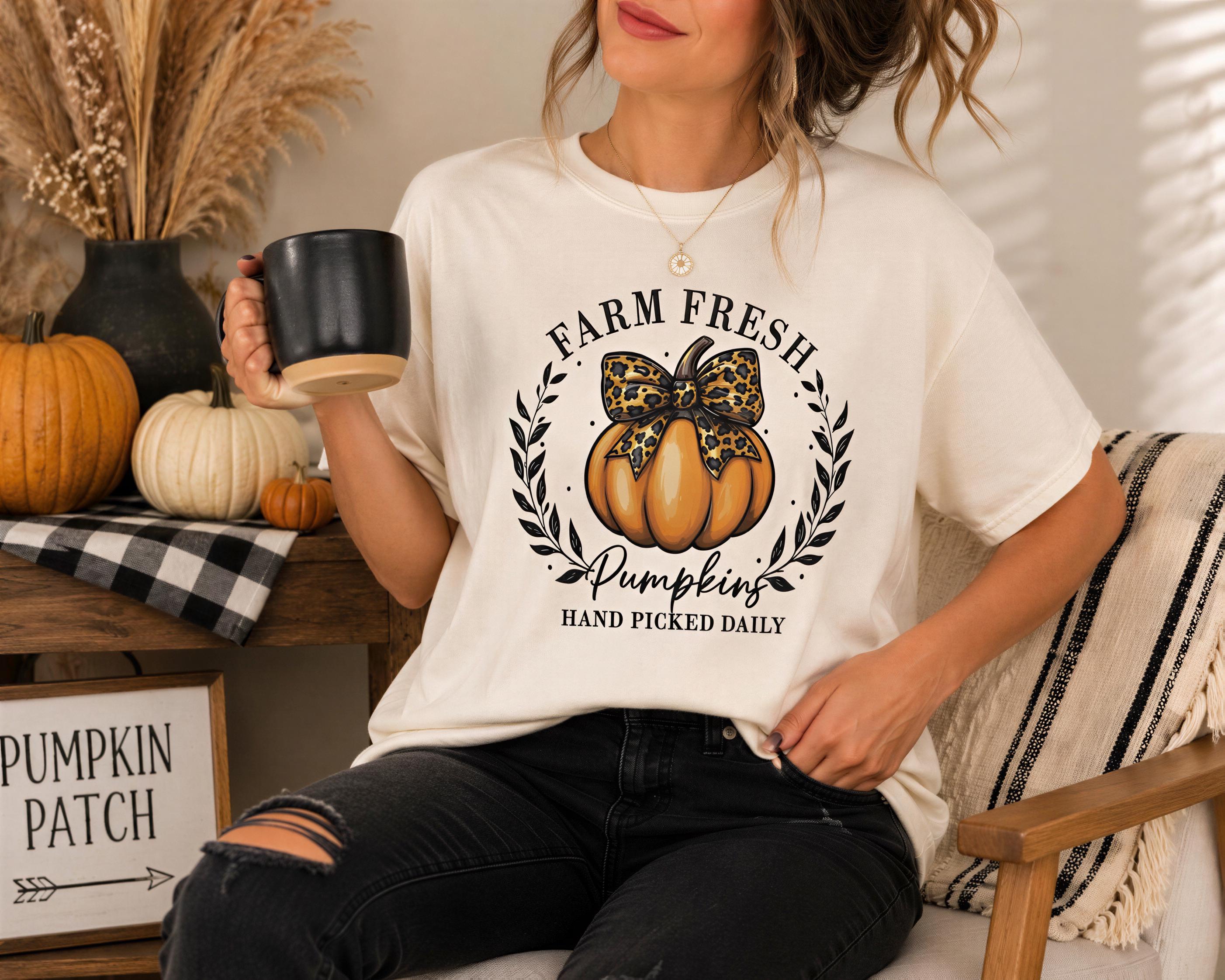 A woman wearing a white t-shirt with a pumpkin graphic and the text "Farm Fresh Pumpkins Hand Picked Daily" is sitting on a chair, holding a black mug. The background includes various fall decorations, such as pumpkins and a plaid blanket.