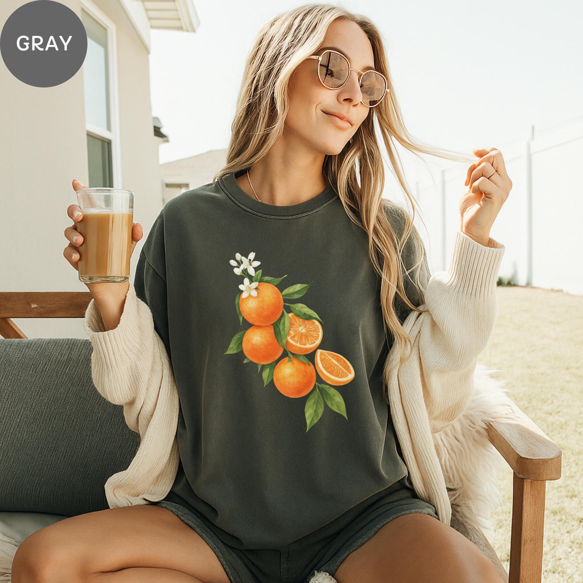 A woman wearing a gray sweatshirt with an orange graphic and holding a glass of orange juice, sitting on a wooden chair outdoors.