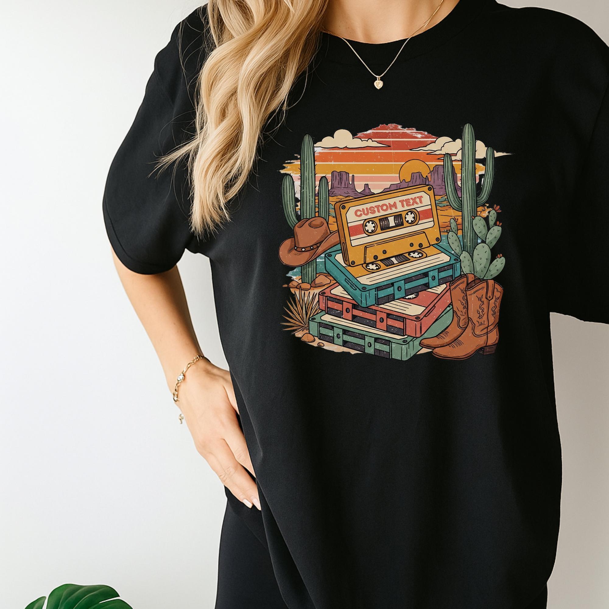 A woman wearing a black t-shirt with a colorful graphic design depicting a cassette tape, cacti, and other desert-themed elements.