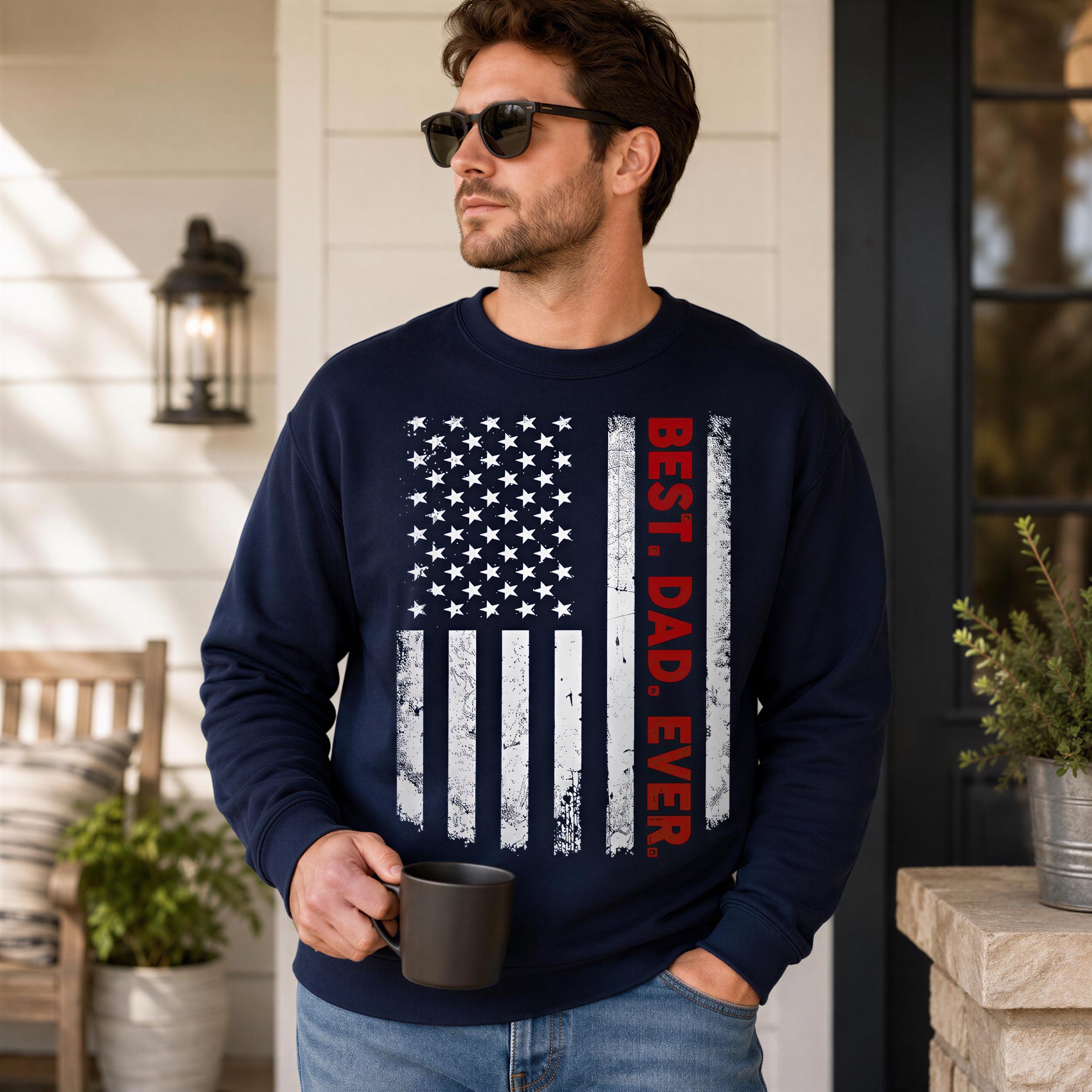 A man wearing a navy blue sweatshirt with an American flag design and the text "BEST. DAD. EVER." is holding a black mug and standing in front of a white house with a porch.