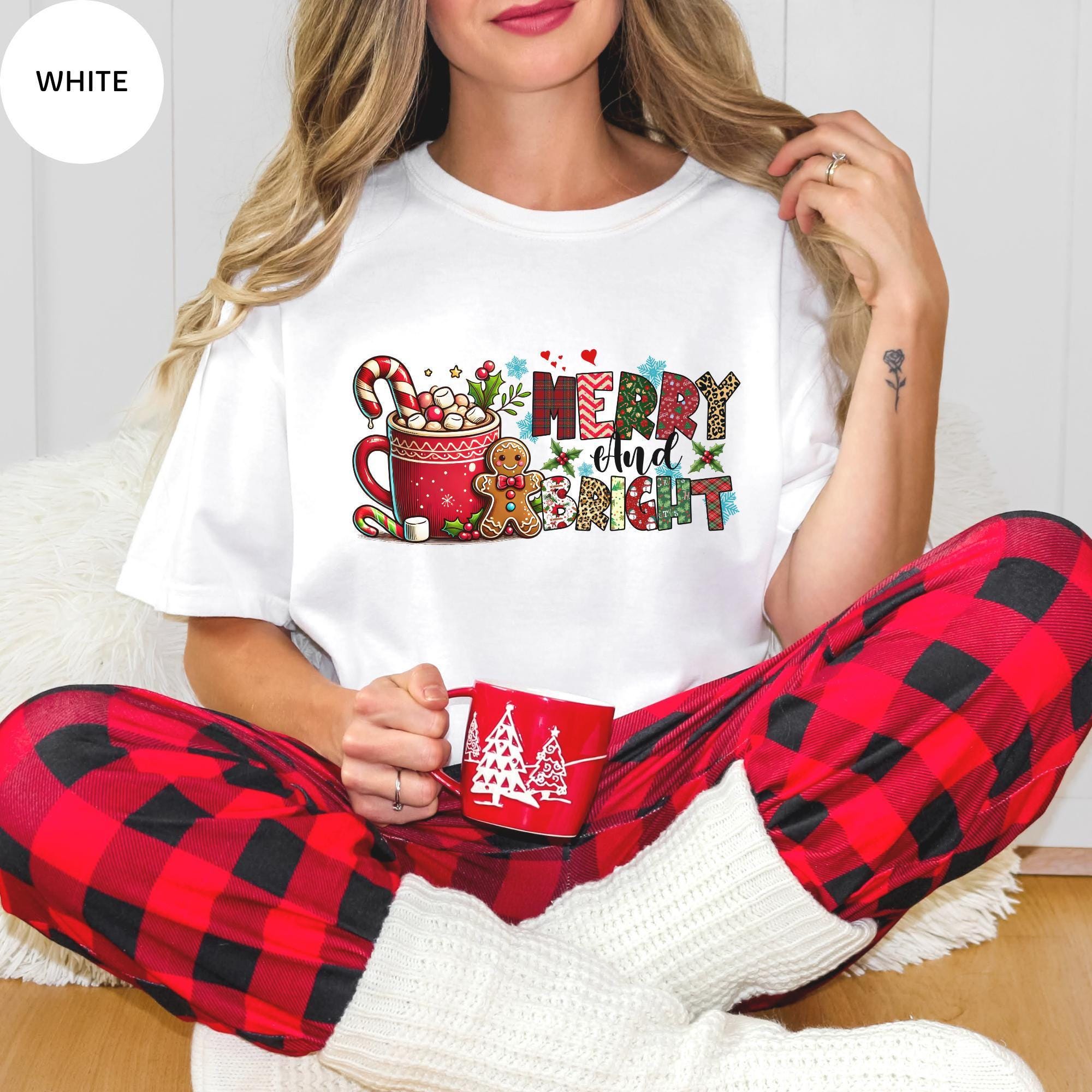 A woman wearing a white t-shirt with a Christmas-themed graphic and red plaid pajama bottoms, holding a red mug.