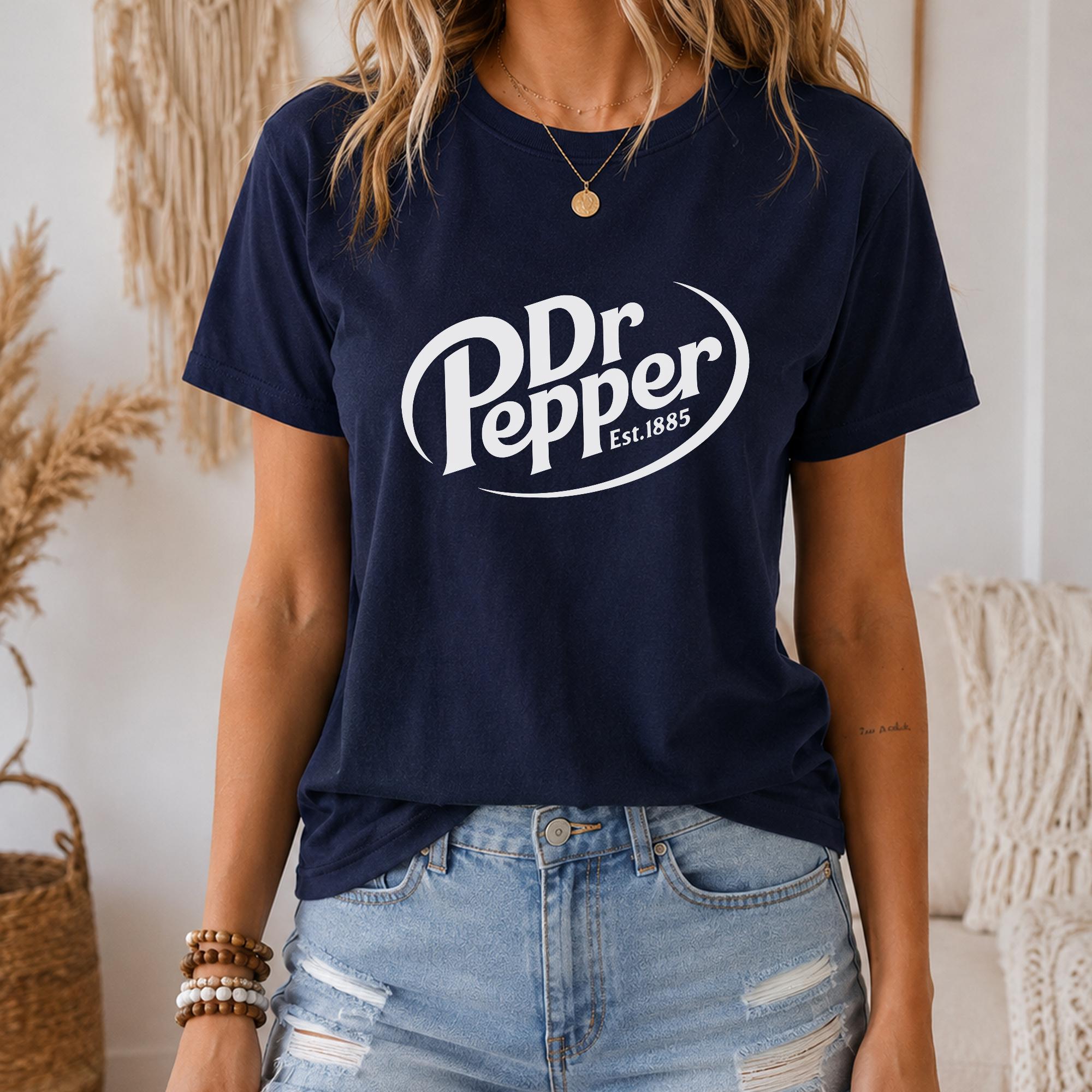 A woman wearing a navy blue t-shirt with the Dr Pepper logo and jeans, standing in a room with a beige wall and a woven basket.