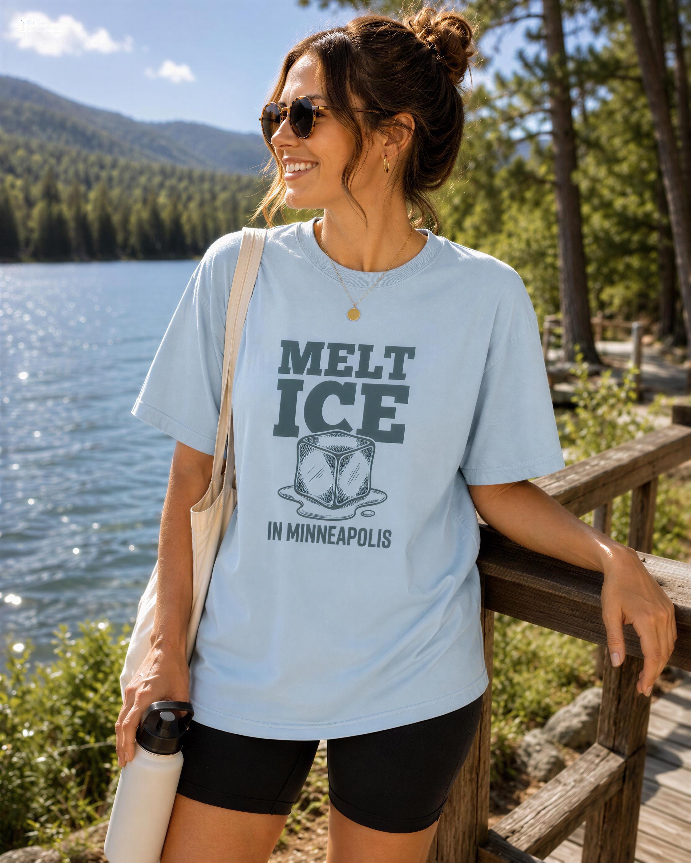 A woman wearing a light blue t-shirt with the text "MELT ICE IN MINNEAPOLIS" standing near a body of water and a wooden fence.