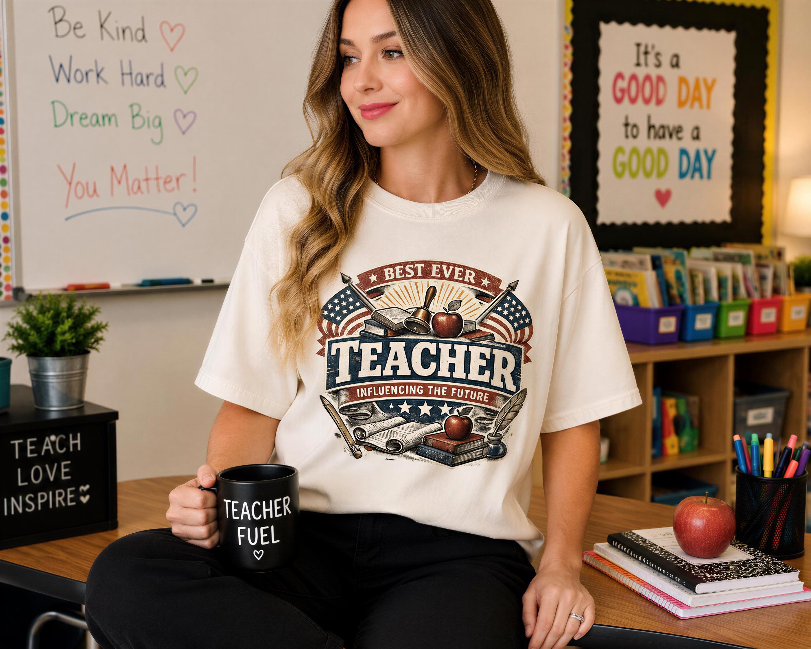 A woman wearing a white t-shirt with a graphic design and holding a black mug with the text "Teacher Fuel" is sitting at a desk in a classroom setting.