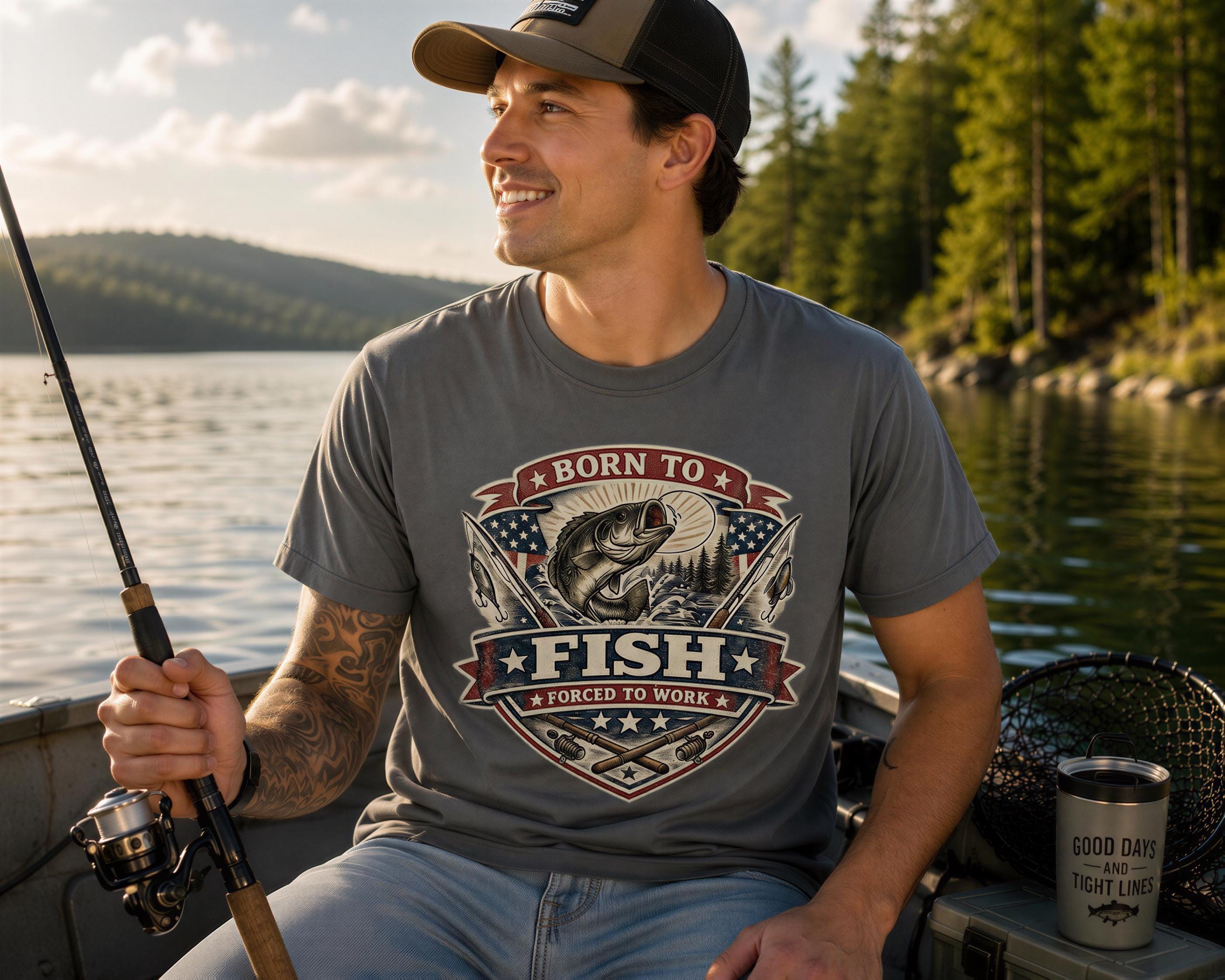 A smiling man wearing a gray t-shirt with a graphic of a fish and the text "Born to fish" is sitting in a boat on a lake, holding a fishing rod.
