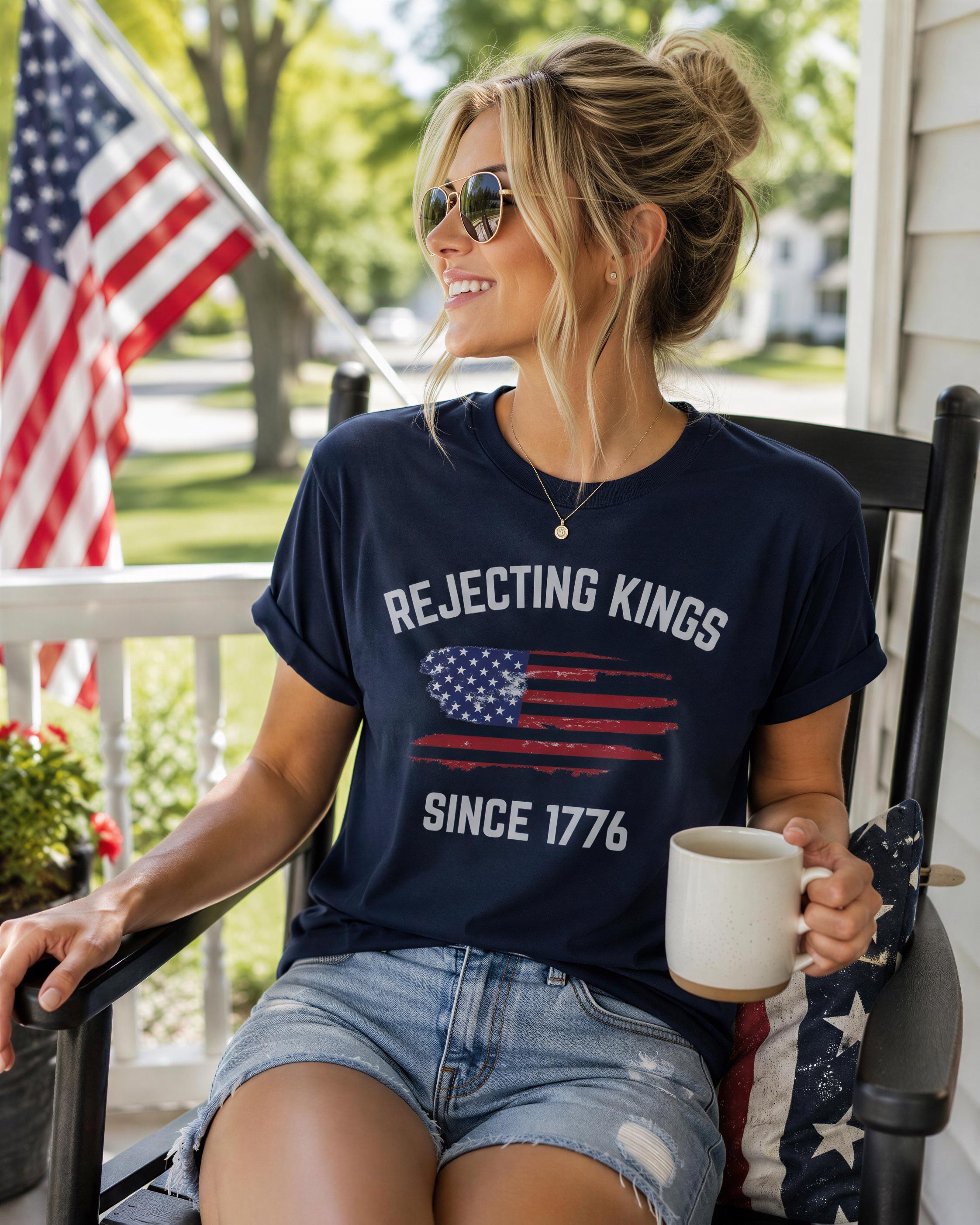 A woman wearing a navy blue t-shirt with the text "REJECTING KINGS SINCE 1776" and sunglasses is sitting on a porch, holding a white mug.