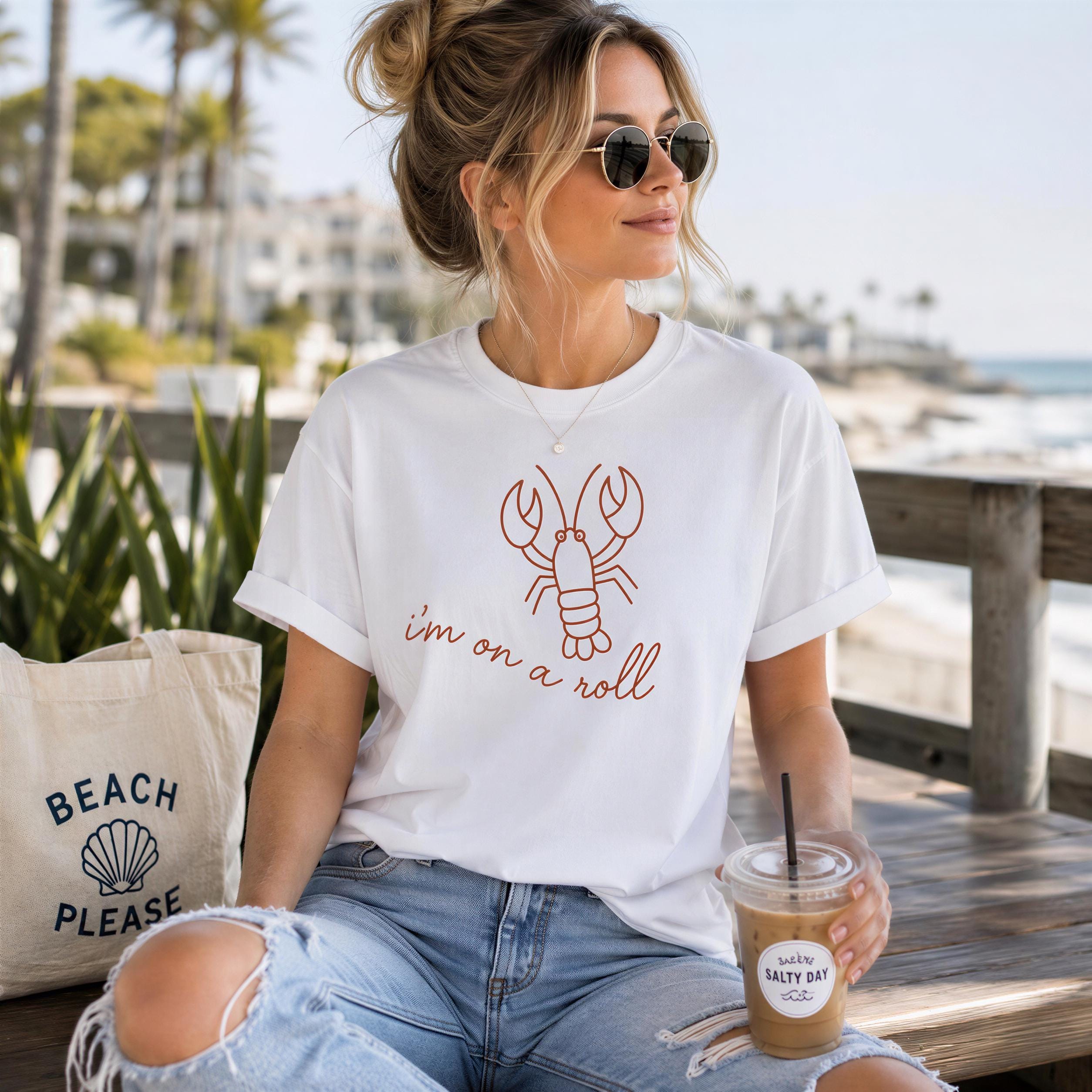 A woman wearing a white t-shirt with a red lobster graphic and sunglasses is sitting on a bench, holding a drink with a straw.