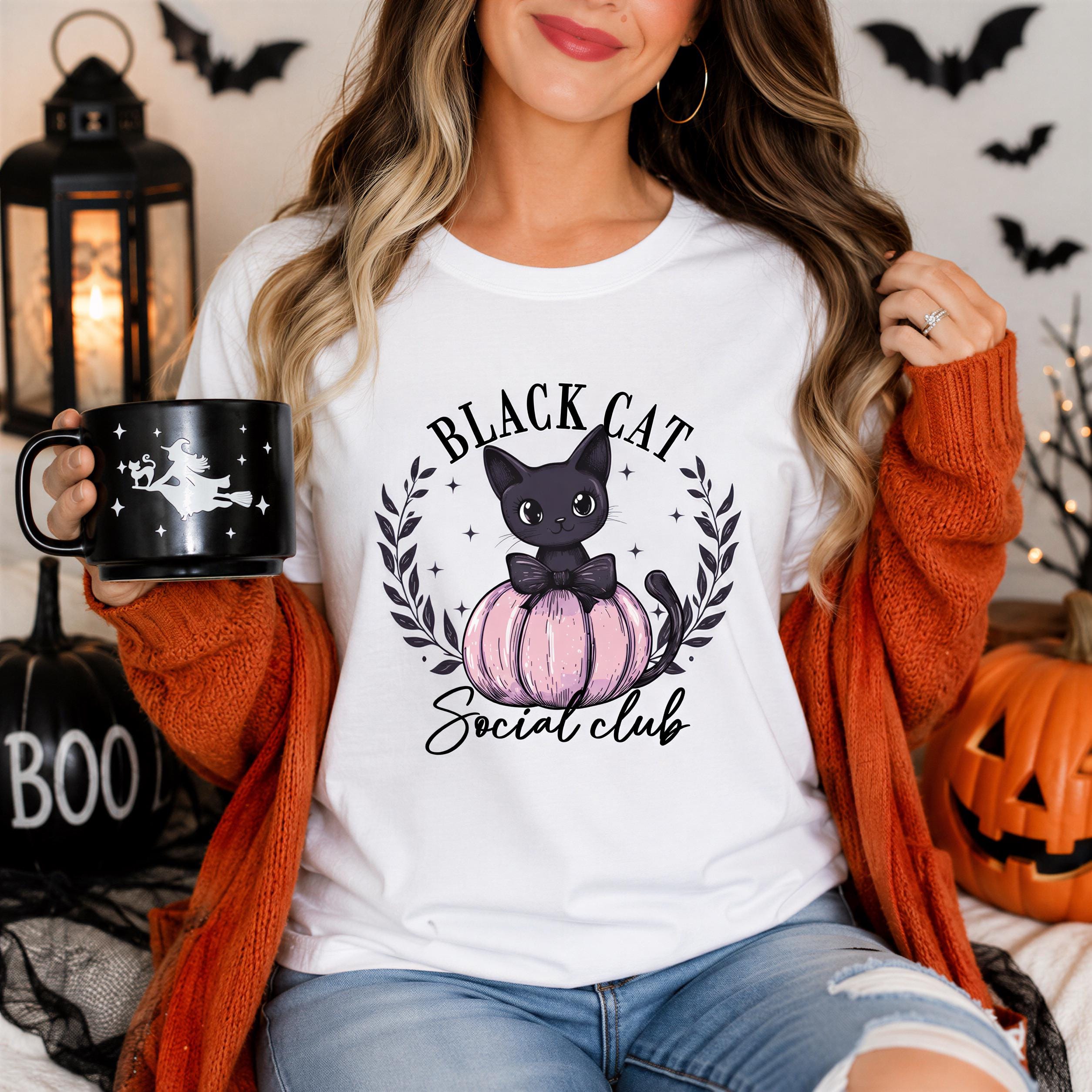 A woman wearing a white t-shirt with a graphic of a cat and pumpkin, holding a black mug with a Halloween-themed design, and surrounded by Halloween decorations.