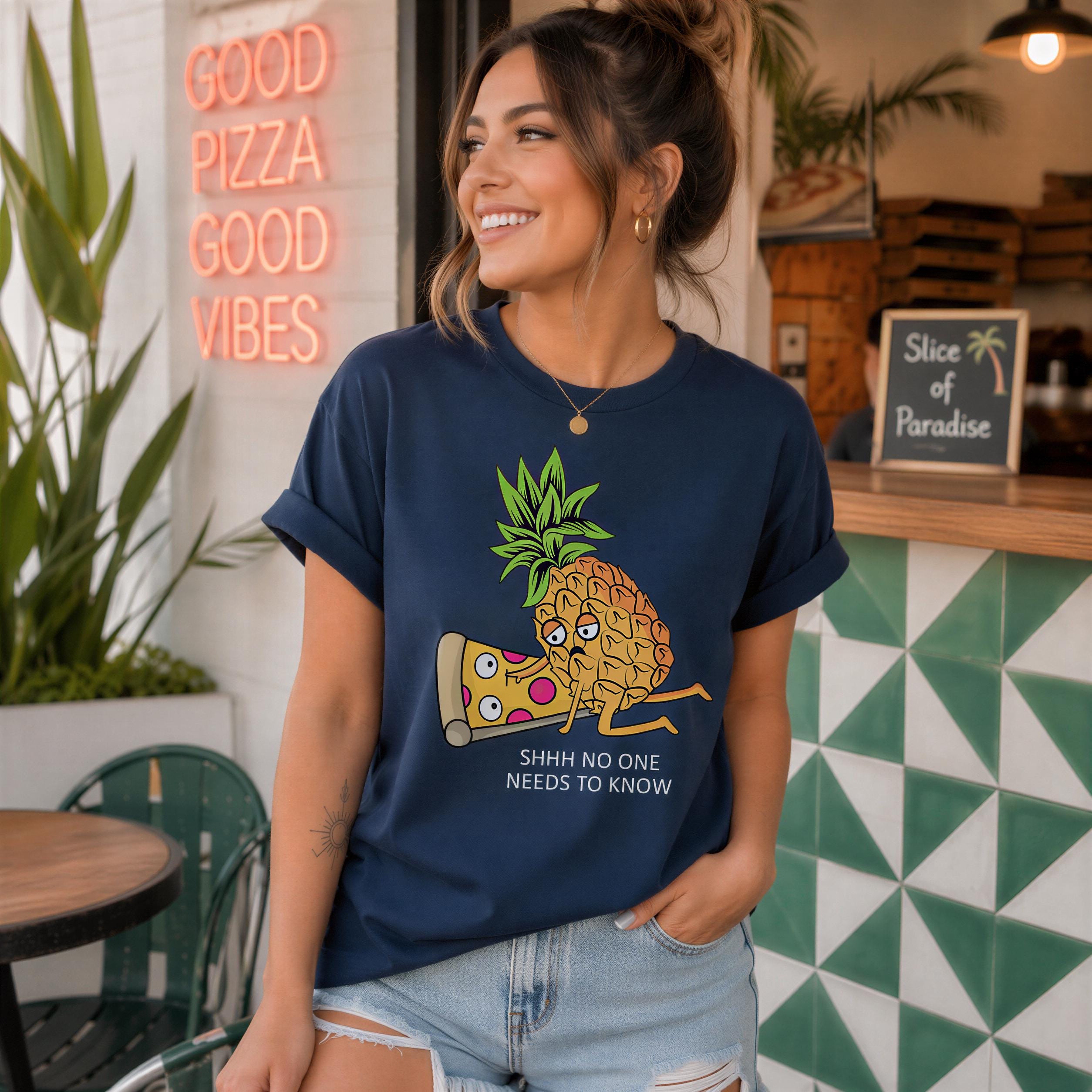 A woman wearing a blue t-shirt with a pineapple graphic and text, smiling and posing in front of a restaurant with a neon sign that says "GOOD PIZZA GOOD VIBES".