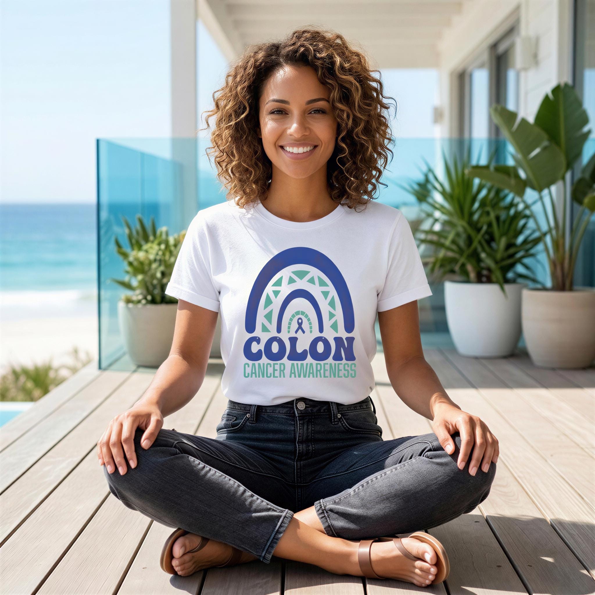 A woman with curly hair is sitting cross-legged on a wooden deck, wearing a white t-shirt with a graphic design and text related to colon cancer awareness.