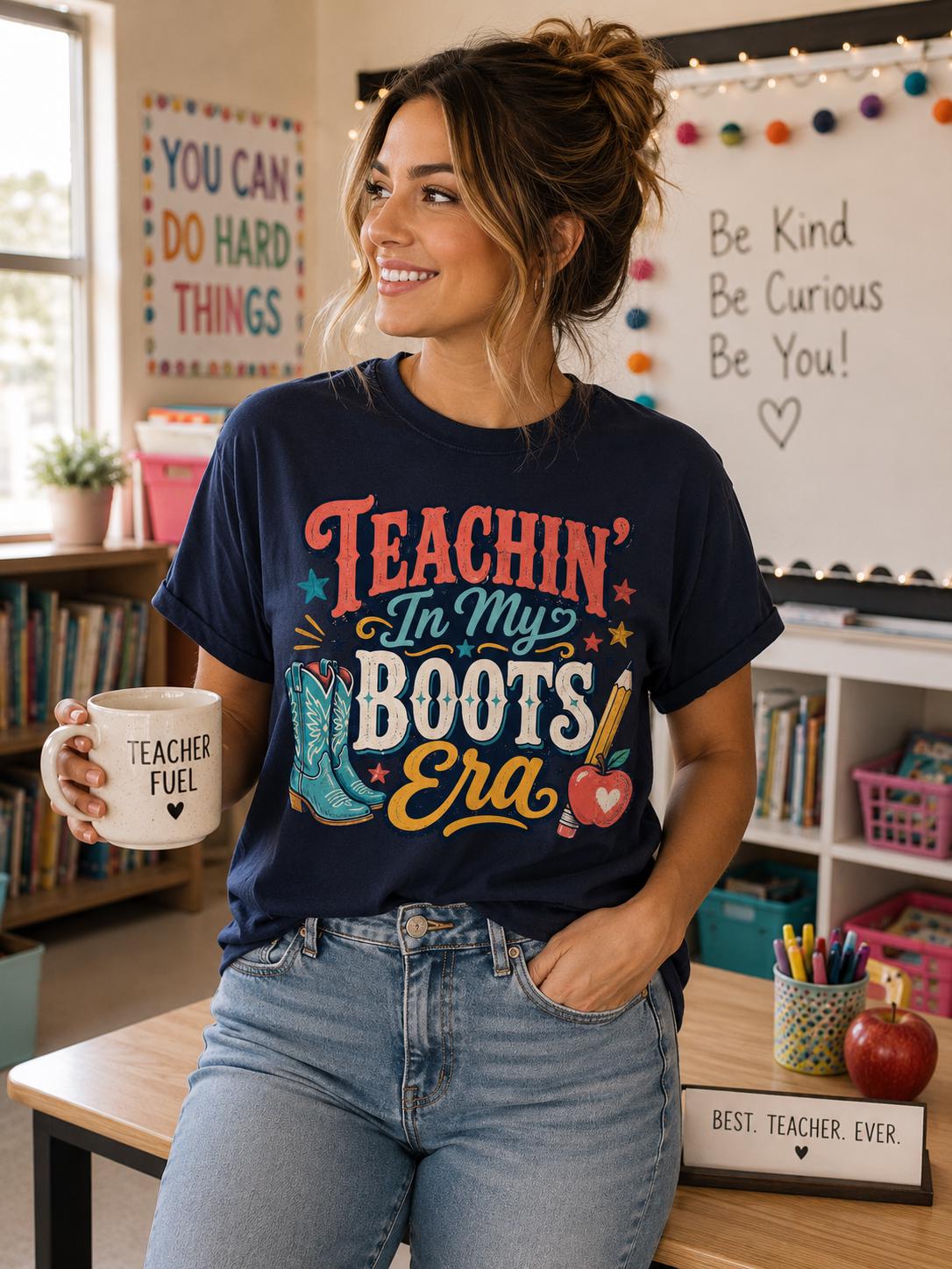 A woman wearing a blue t-shirt with a colorful design and holding a coffee mug stands in a classroom with a bulletin board and various school supplies visible in the background.
