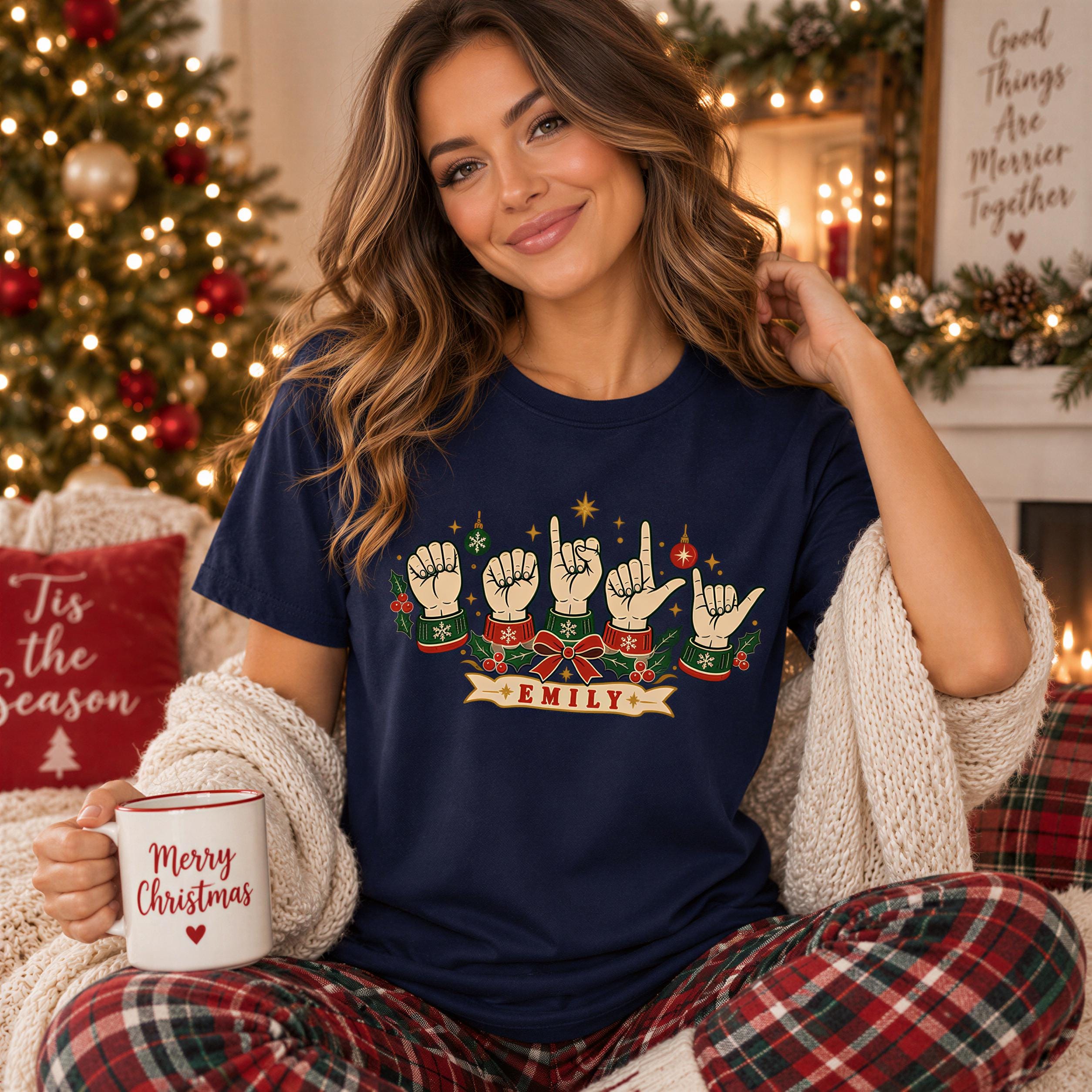 A woman wearing a navy blue t-shirt with a graphic of hand gestures and the name "EMILY" is sitting in a cozy setting with a Christmas tree and other holiday decorations in the background.