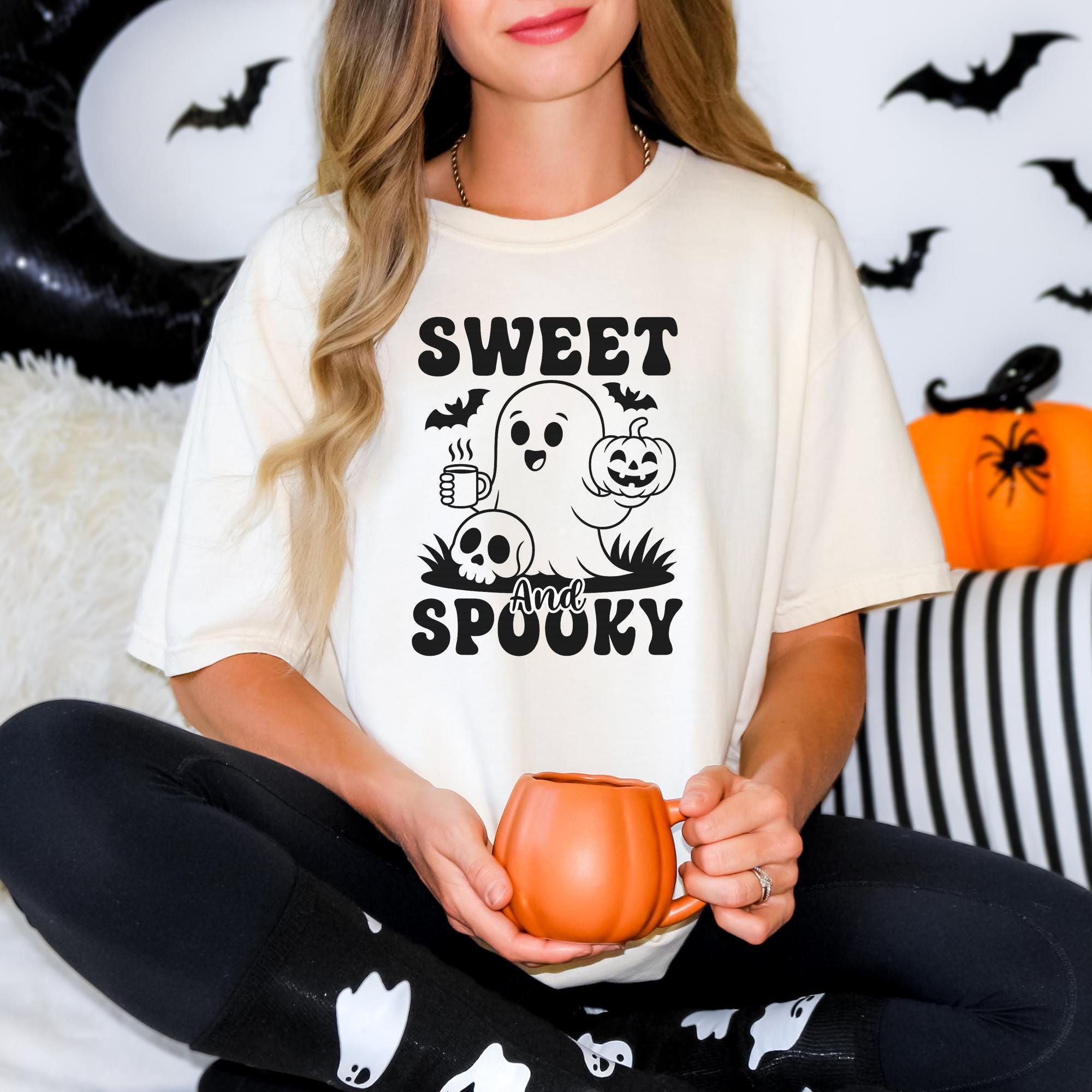 A woman wearing a white t-shirt with a graphic design of a ghost holding a pumpkin and a cup, sitting on a couch with Halloween decorations in the background.