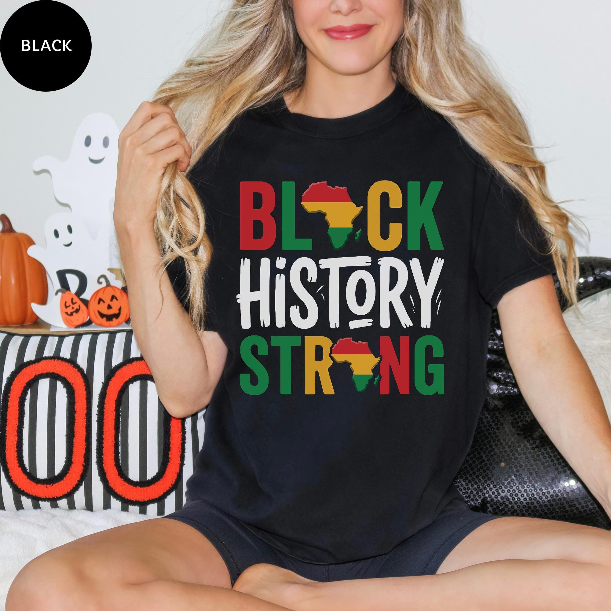 A woman wearing a black t-shirt with the text "BLACK HISTORY STRONG" printed on it, sitting on a couch.