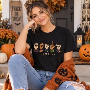 Könnte beinhalten: Ein schwarzes T-Shirt mit einer Grafik von fünf Händen in amerikanischer Gebärdensprache, die den Namen EMILY buchstabieren. Jede Hand ist mit bunten Halloween-Manschetten verziert, darunter Muster mit Fledermäusen, Streifen, Spinnennetzen und Sternen.