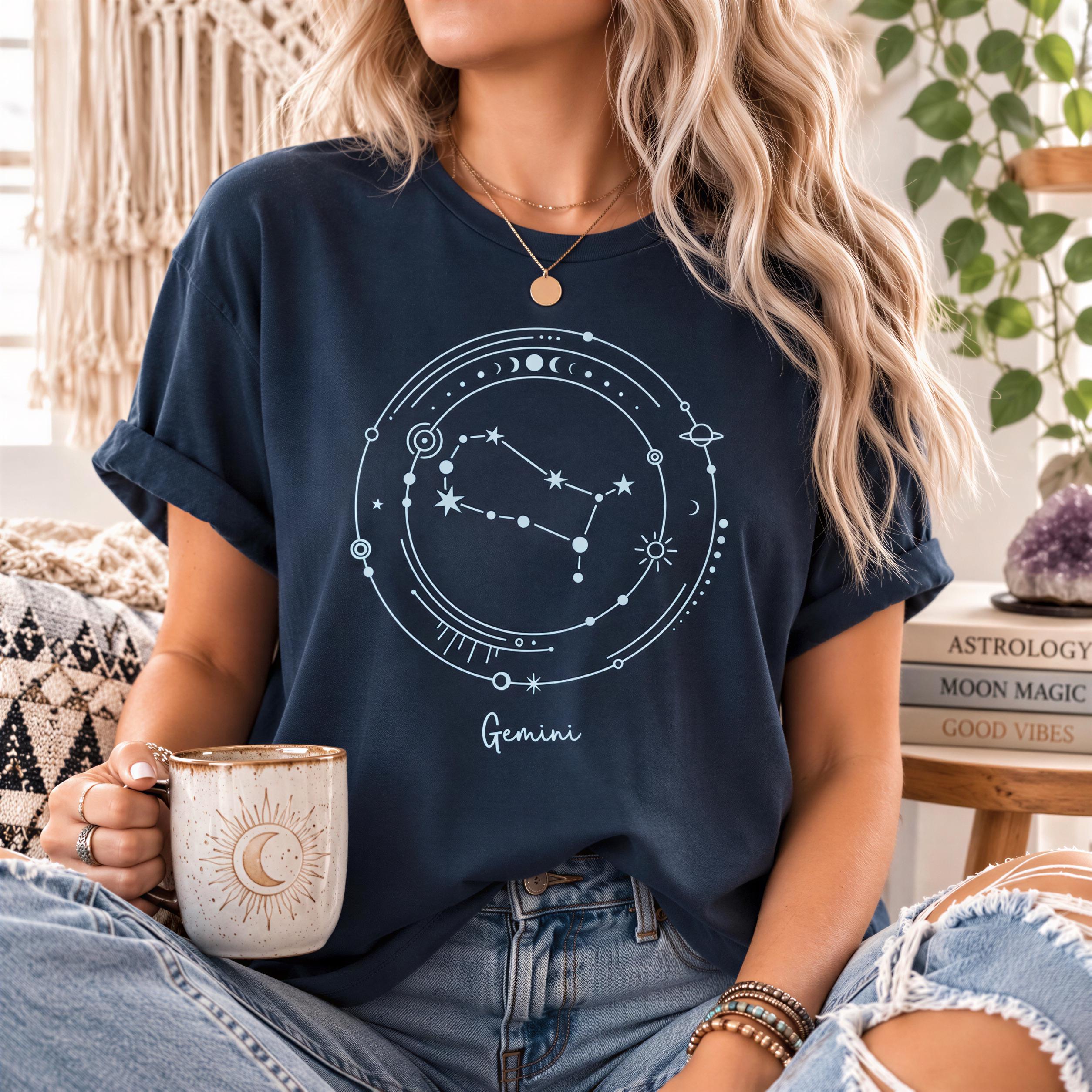 A woman wearing a navy blue t-shirt with a graphic of the zodiac sign Gemini, holding a coffee mug, and sitting on a couch with a potted plant in the background.