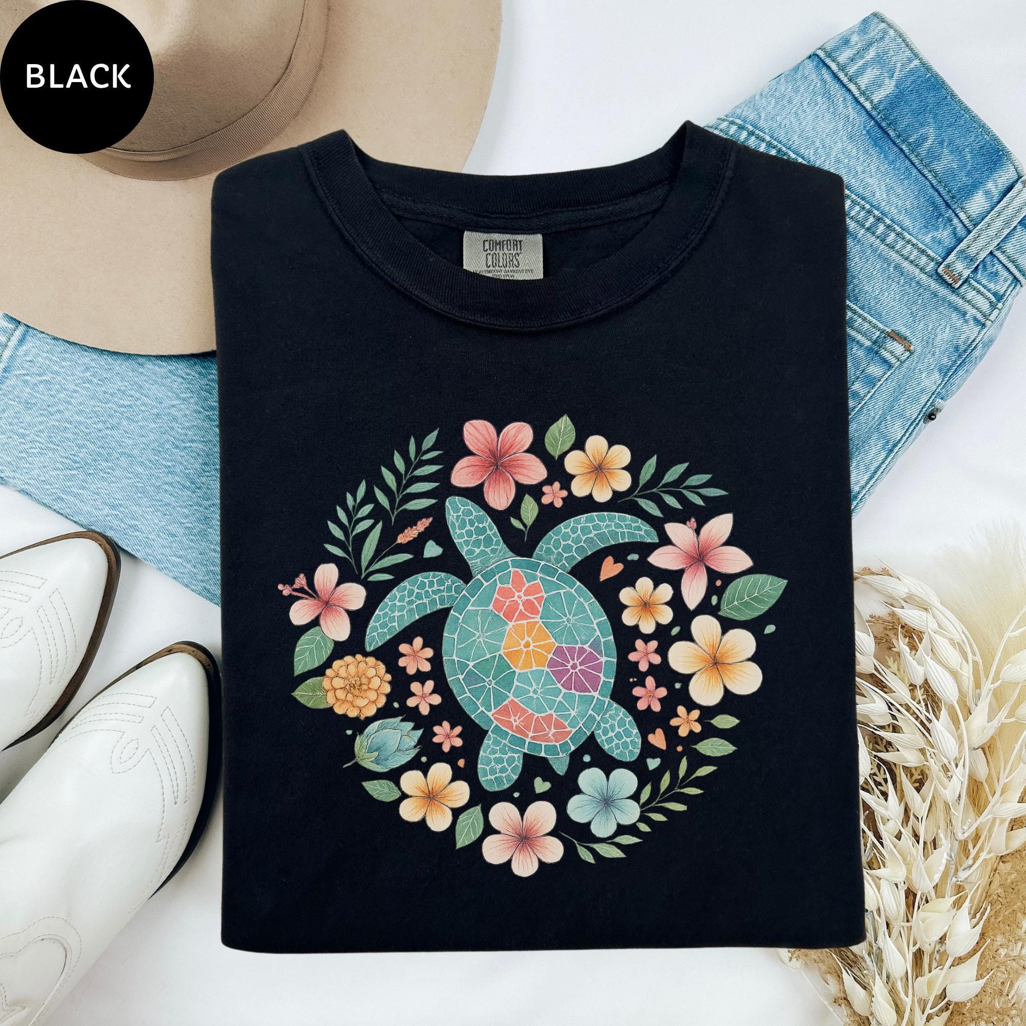 a black t-shirt with a colorful floral design, including a turtle surrounded by flowers and leaves. The t-shirt is placed on a white surface, with a pair of white boots and a hat nearby.