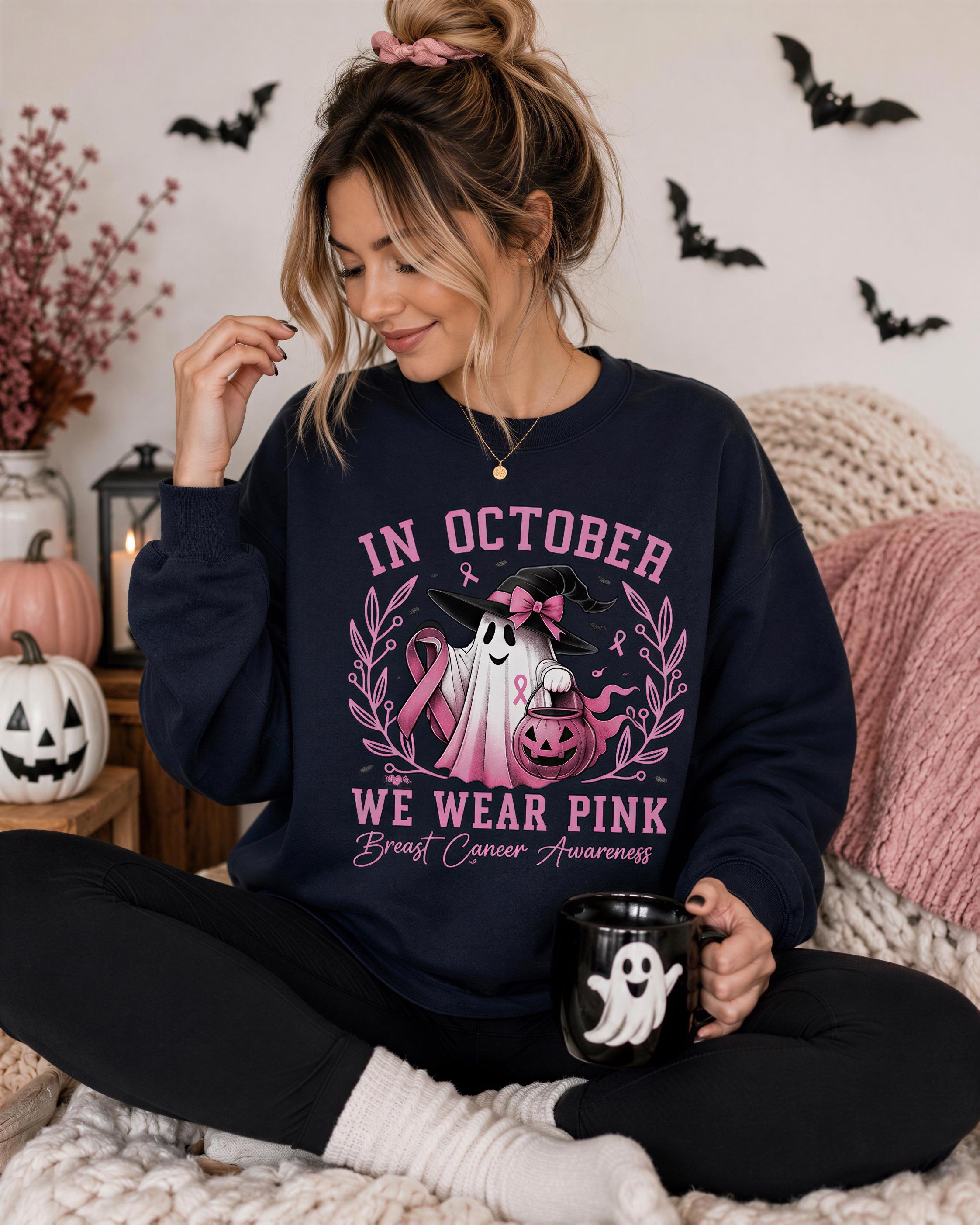 A woman wearing a black sweatshirt with a graphic of a ghost and a pink ribbon, sitting on a bed with Halloween decorations in the background.