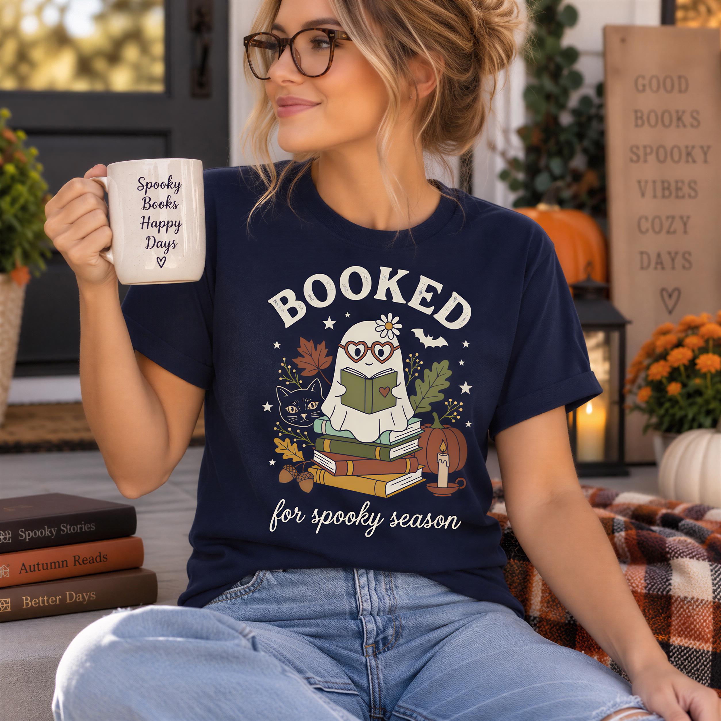A woman wearing a navy blue t-shirt with a graphic of a ghost reading a book and the text "BOOKED for spooky season" is sitting on a couch with a mug in her hand.