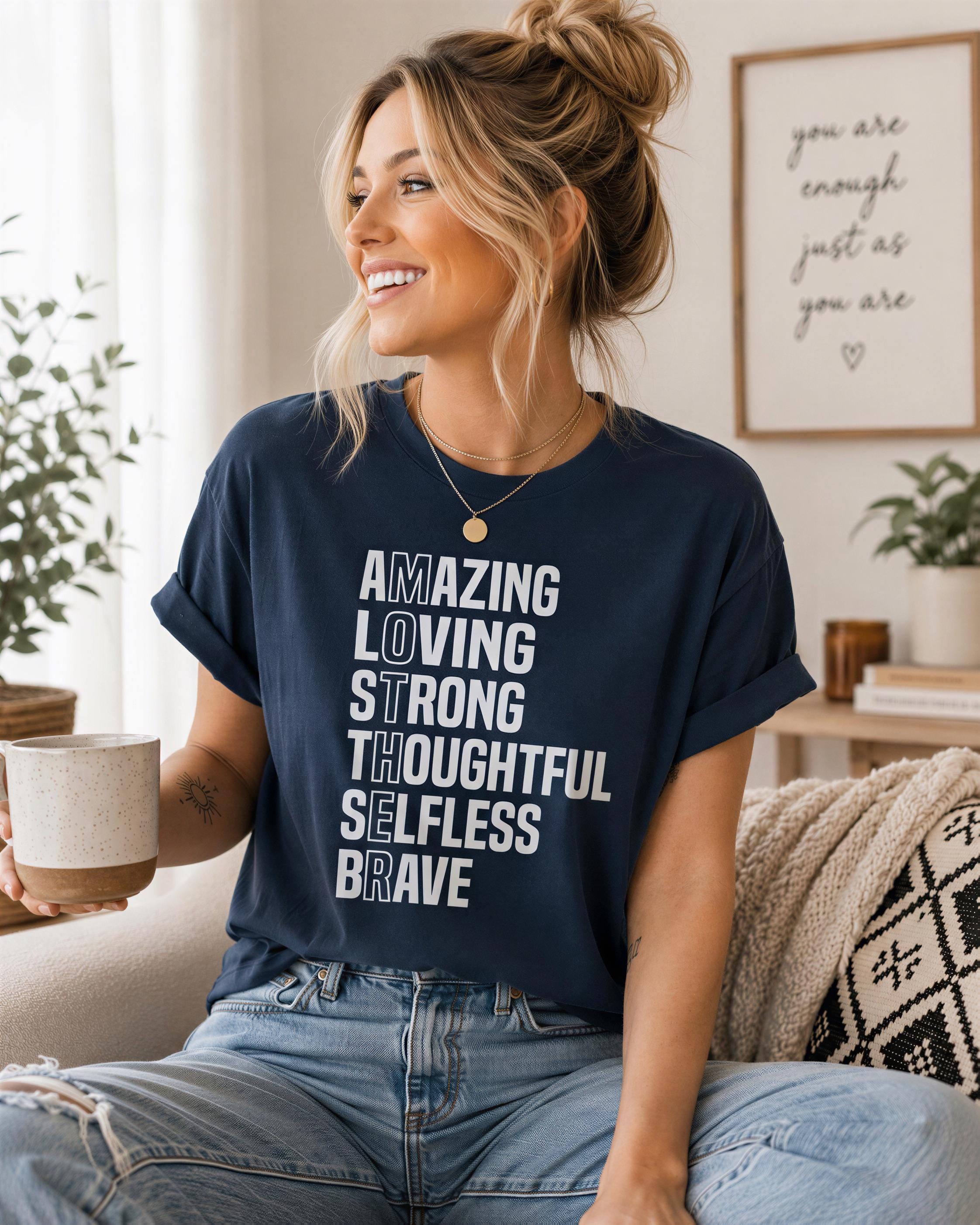 A woman wearing a navy blue t-shirt with white text and a gold necklace is sitting on a couch, smiling and holding a coffee mug.