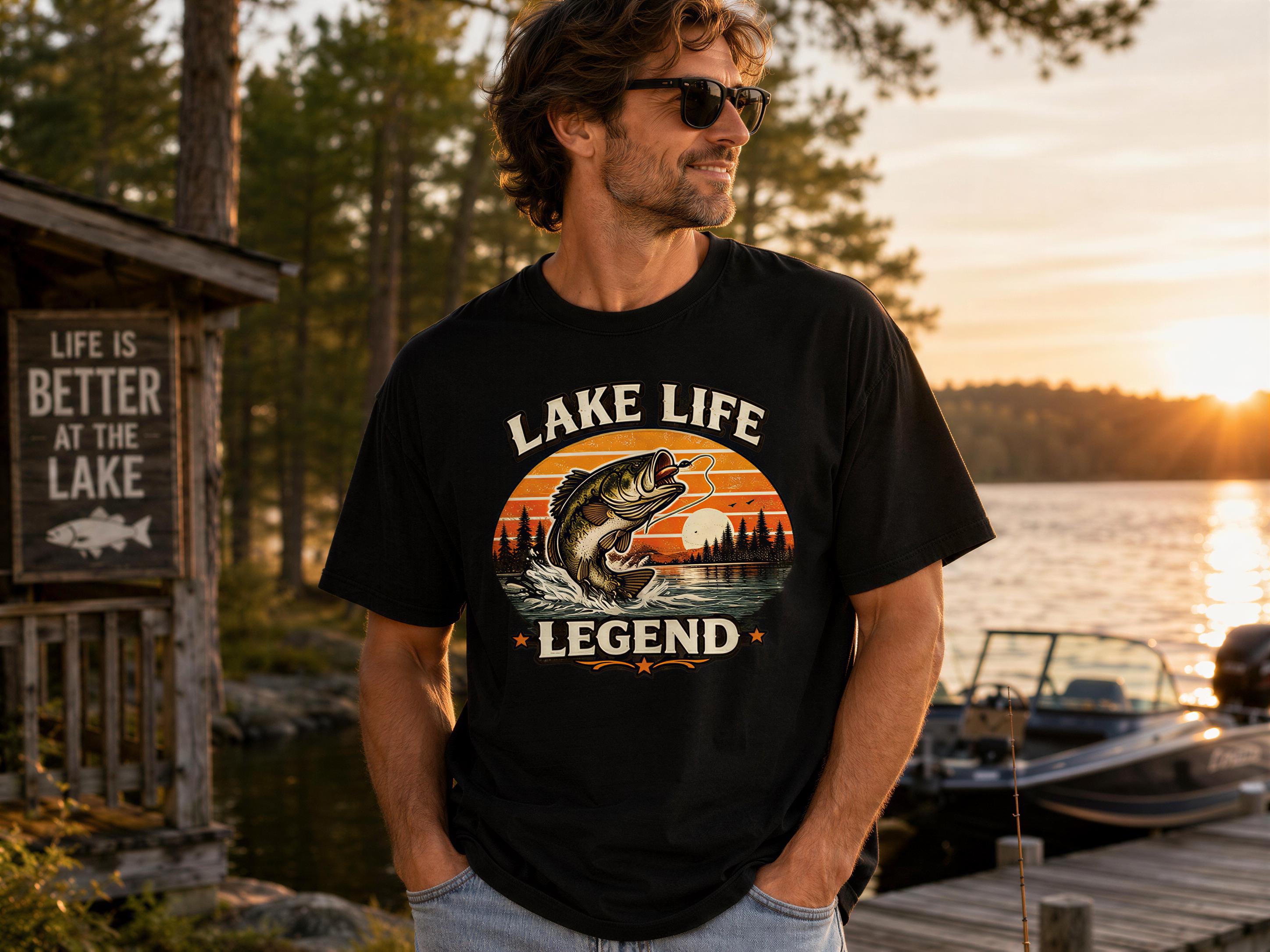 A man wearing a black t-shirt with a graphic of a fish and the text "LAKELIFE LEGEND" stands near a lake with a wooden dock and a sign that says "LIFE IS BETTER AT THE LAKE".