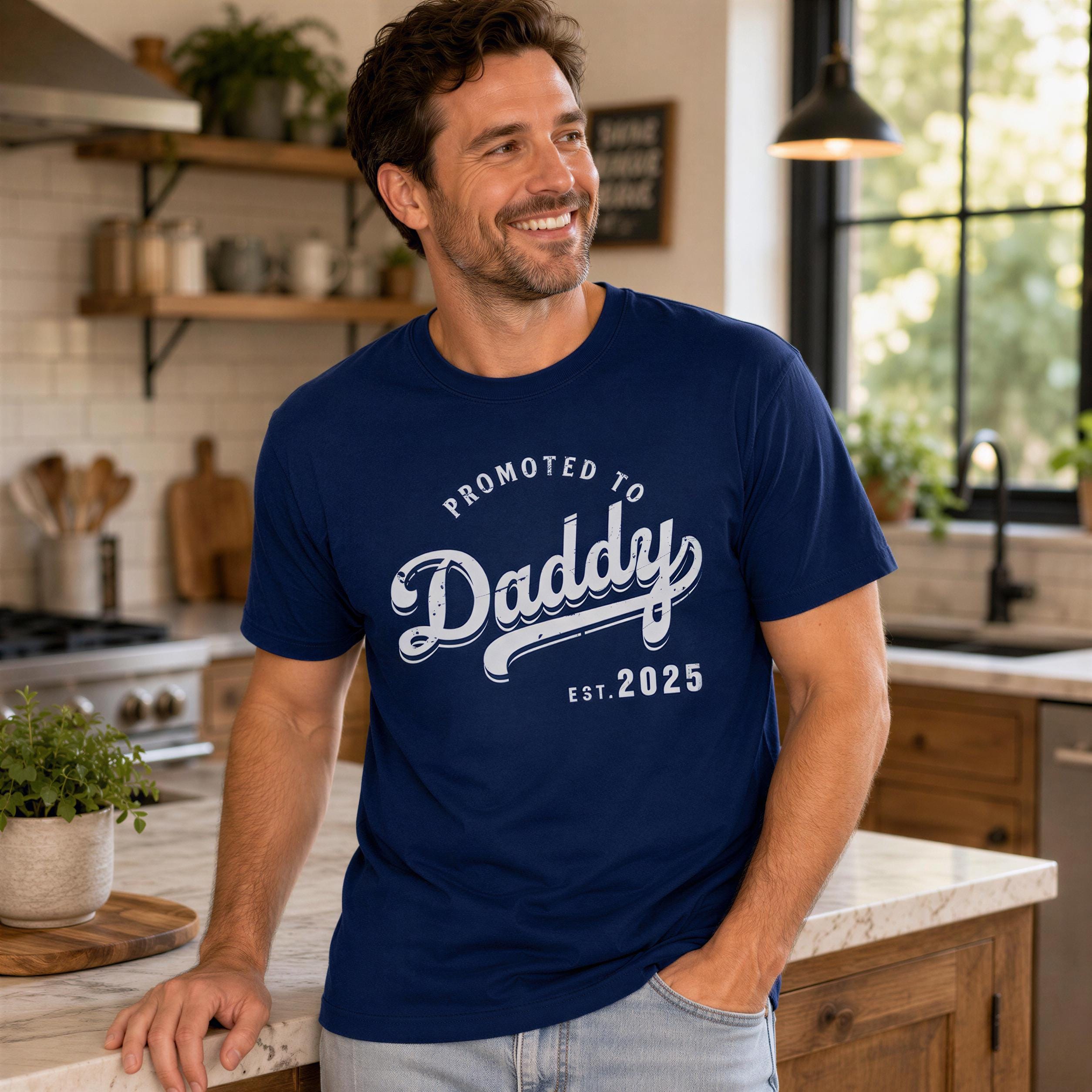A man wearing a blue t-shirt with the text "Promoted to Daddy" is standing in a kitchen, smiling.