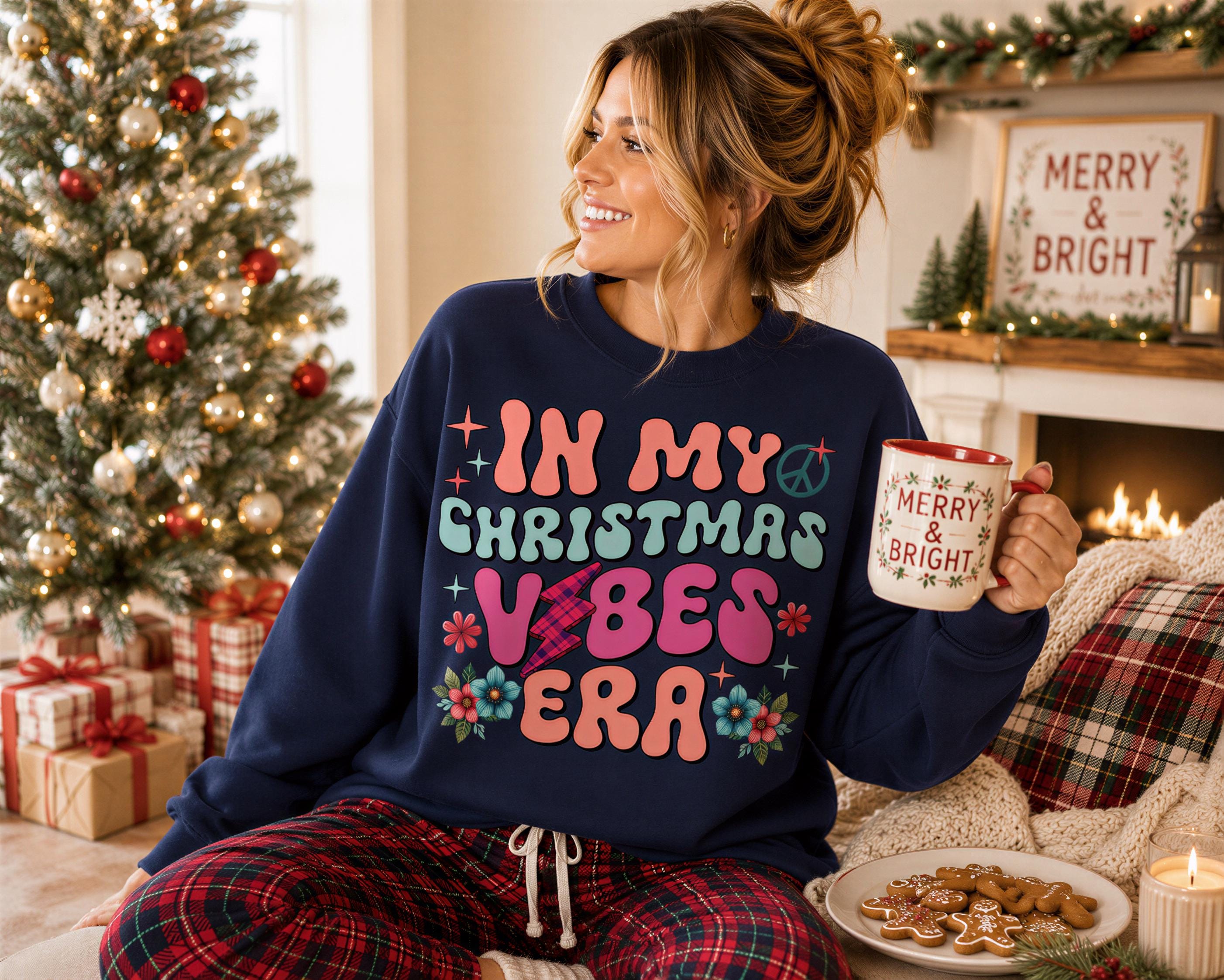 A woman wearing a blue sweatshirt with a colorful design and holding a mug is sitting in front of a Christmas tree and a fireplace.