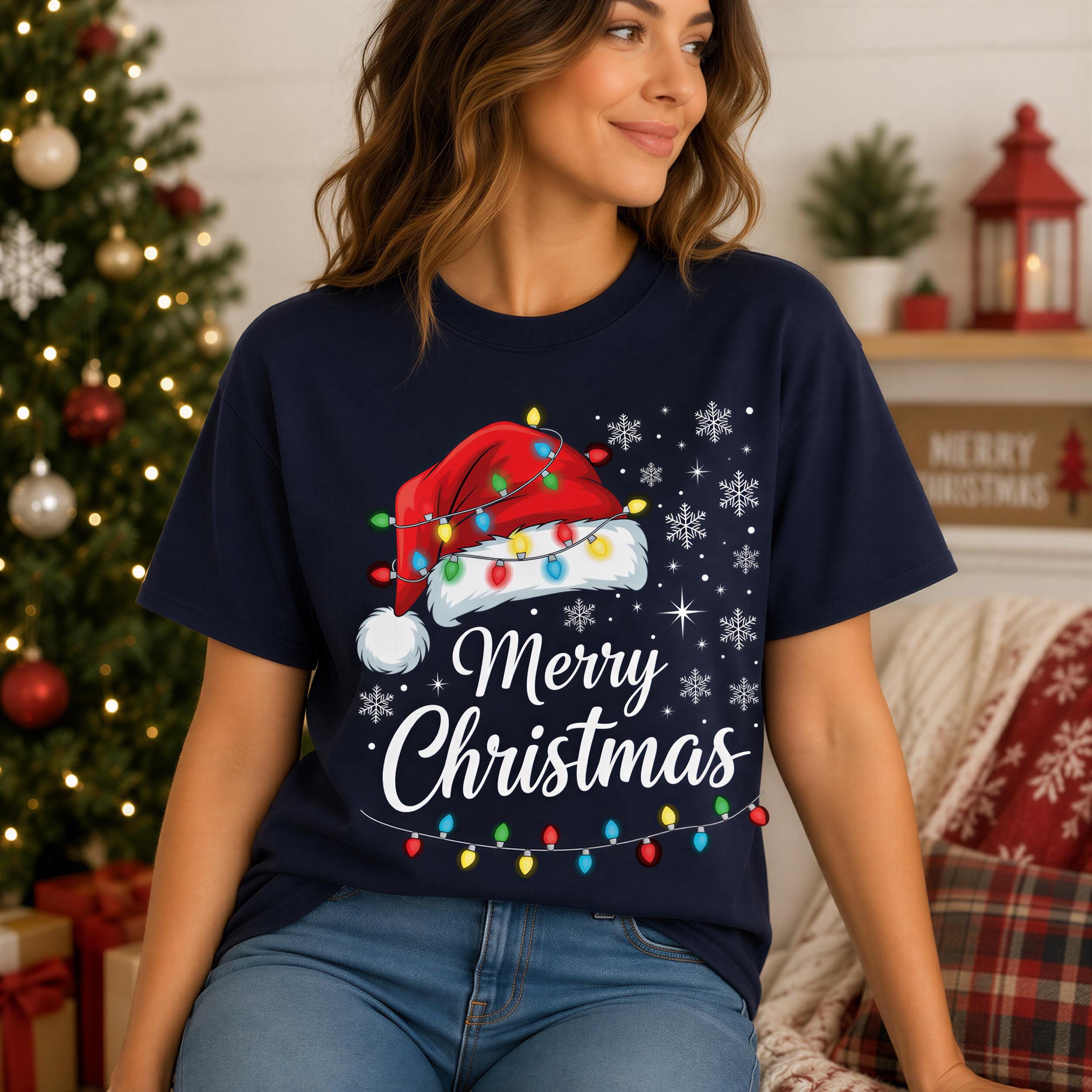 A woman wearing a navy blue t-shirt with a Christmas-themed design, including a Santa hat and snowflakes, is sitting in front of a Christmas tree.