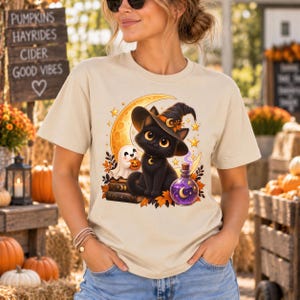 May include: A beige t-shirt featuring a graphic of a black cat wearing a witch hat, sitting on books next to a small ghost holding a pumpkin. The design includes a yellow crescent moon, stars, autumn leaves, and a purple potion bottle.