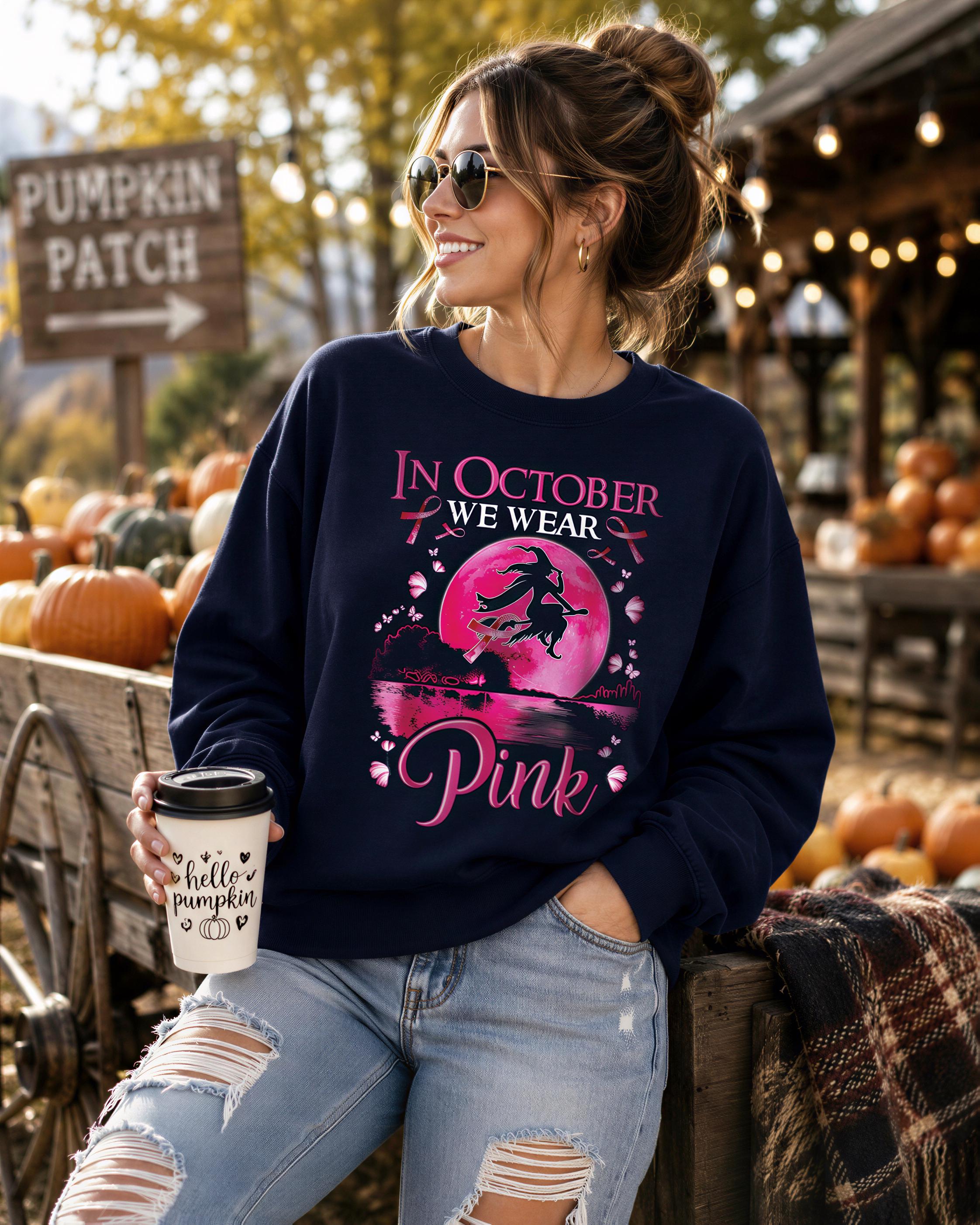 A woman wearing a navy blue sweatshirt with a pink design and text, holding a coffee cup, standing in front of a pumpkin patch with a sign that says 'PUMPKIN PATCH'.