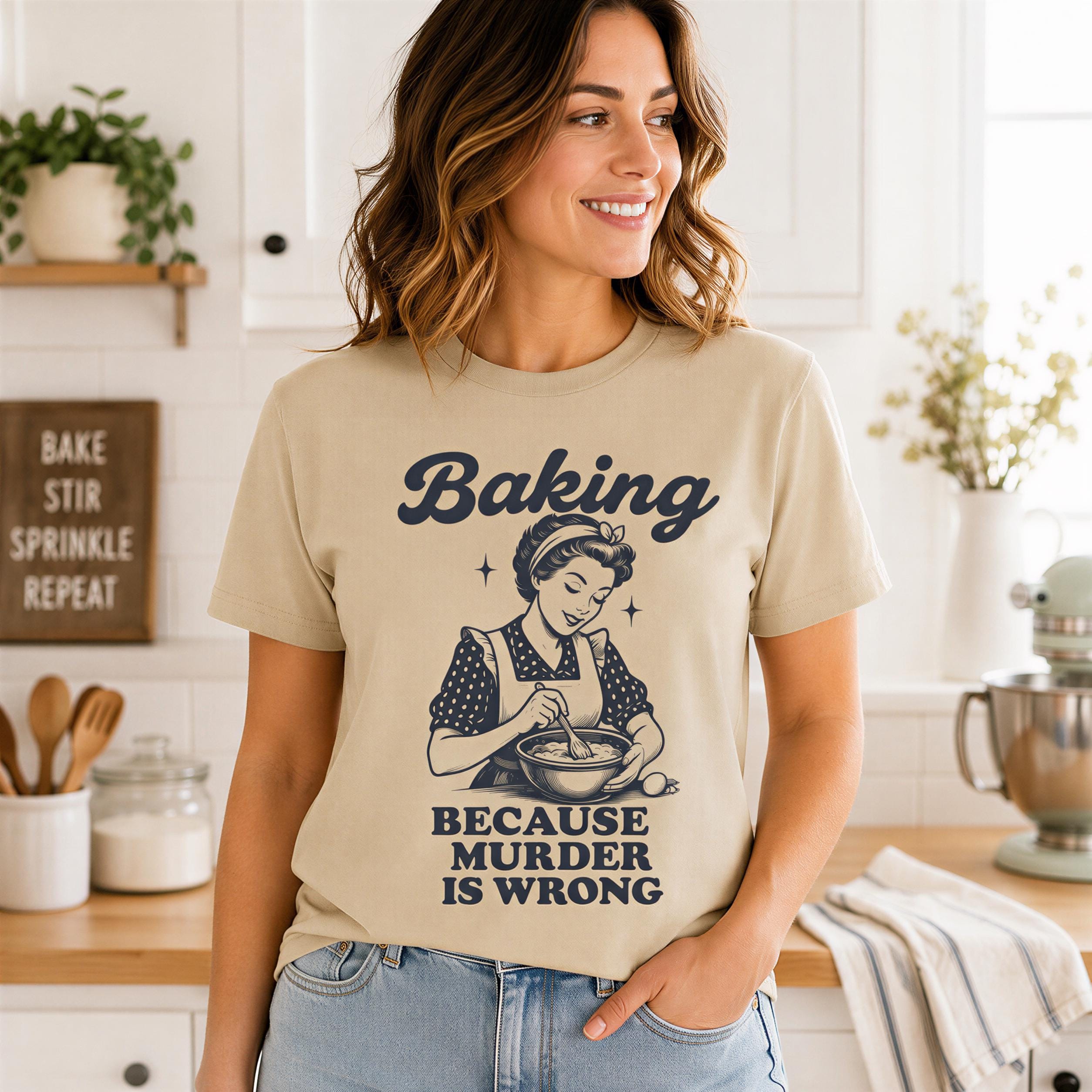A woman wearing a t-shirt with a humorous graphic and text, standing in a kitchen with various kitchen items visible in the background.