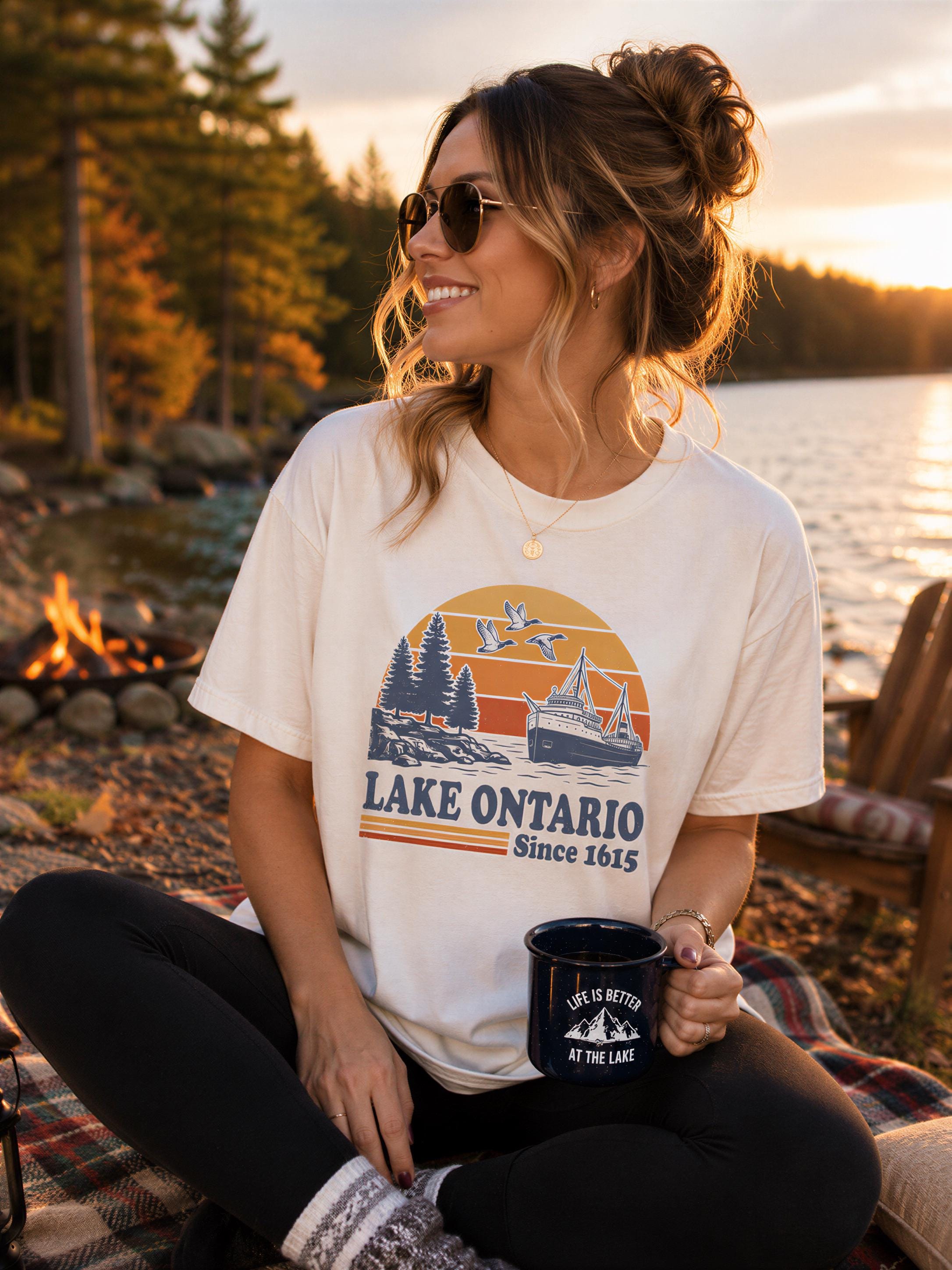 A woman sitting on a blanket by a lake, wearing a white t-shirt with a graphic design and holding a mug, with a sunset in the background.