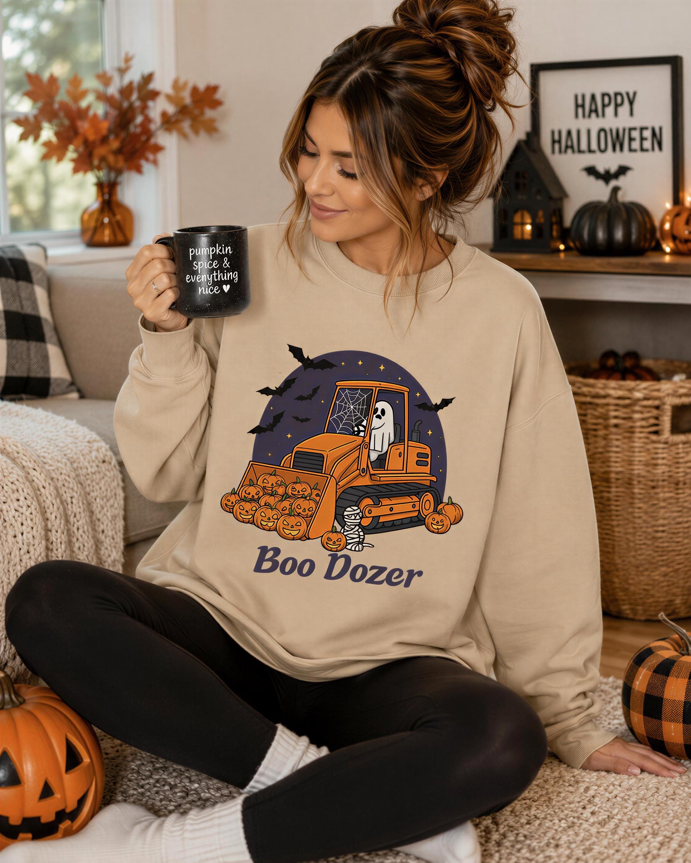 A woman sitting on the floor, wearing a beige sweatshirt with a Halloween-themed graphic, holding a mug and surrounded by Halloween decorations.