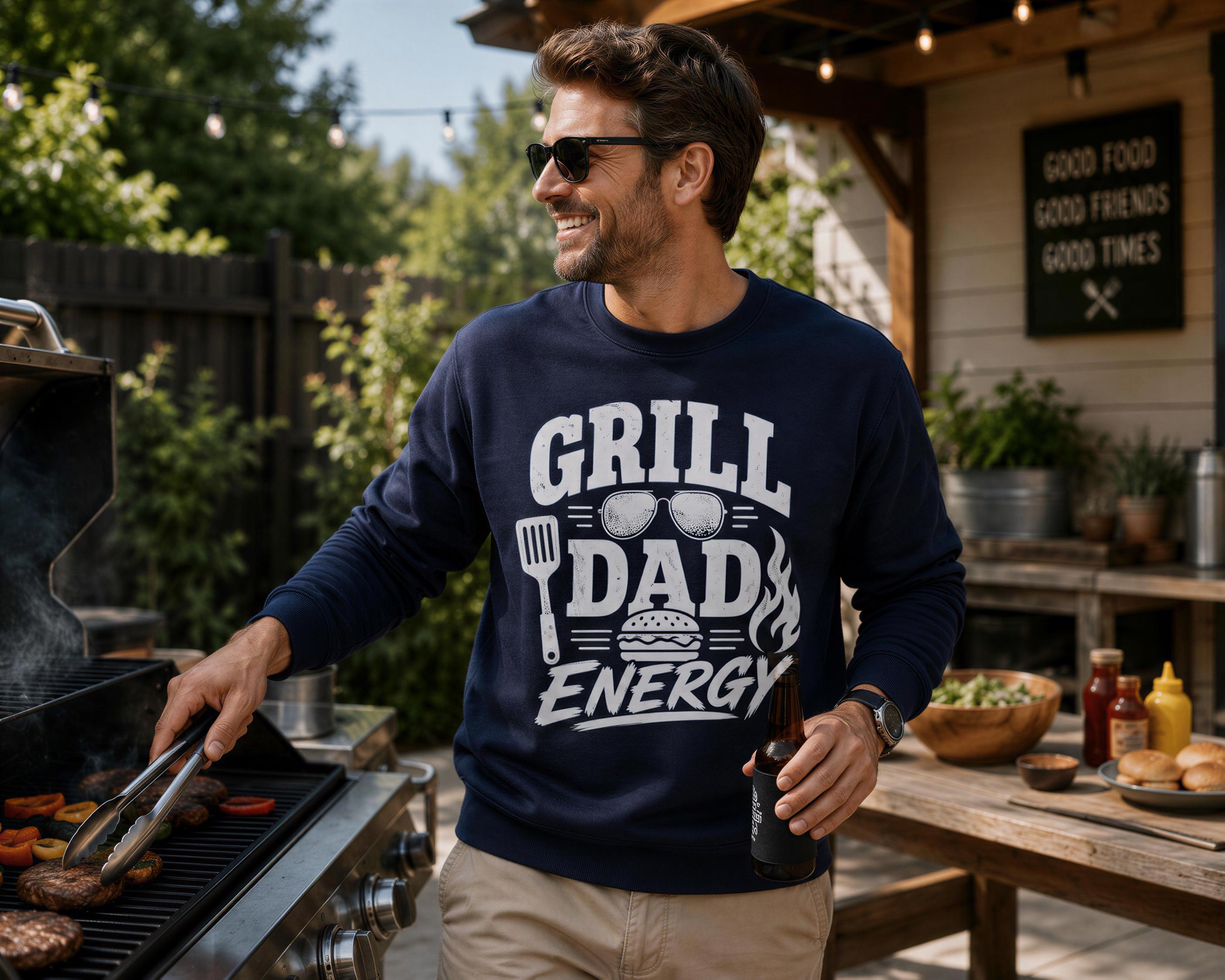 A man in a blue sweatshirt with the text "Grill Dad Energy" is grilling food outdoors, smiling and holding a beer bottle.
