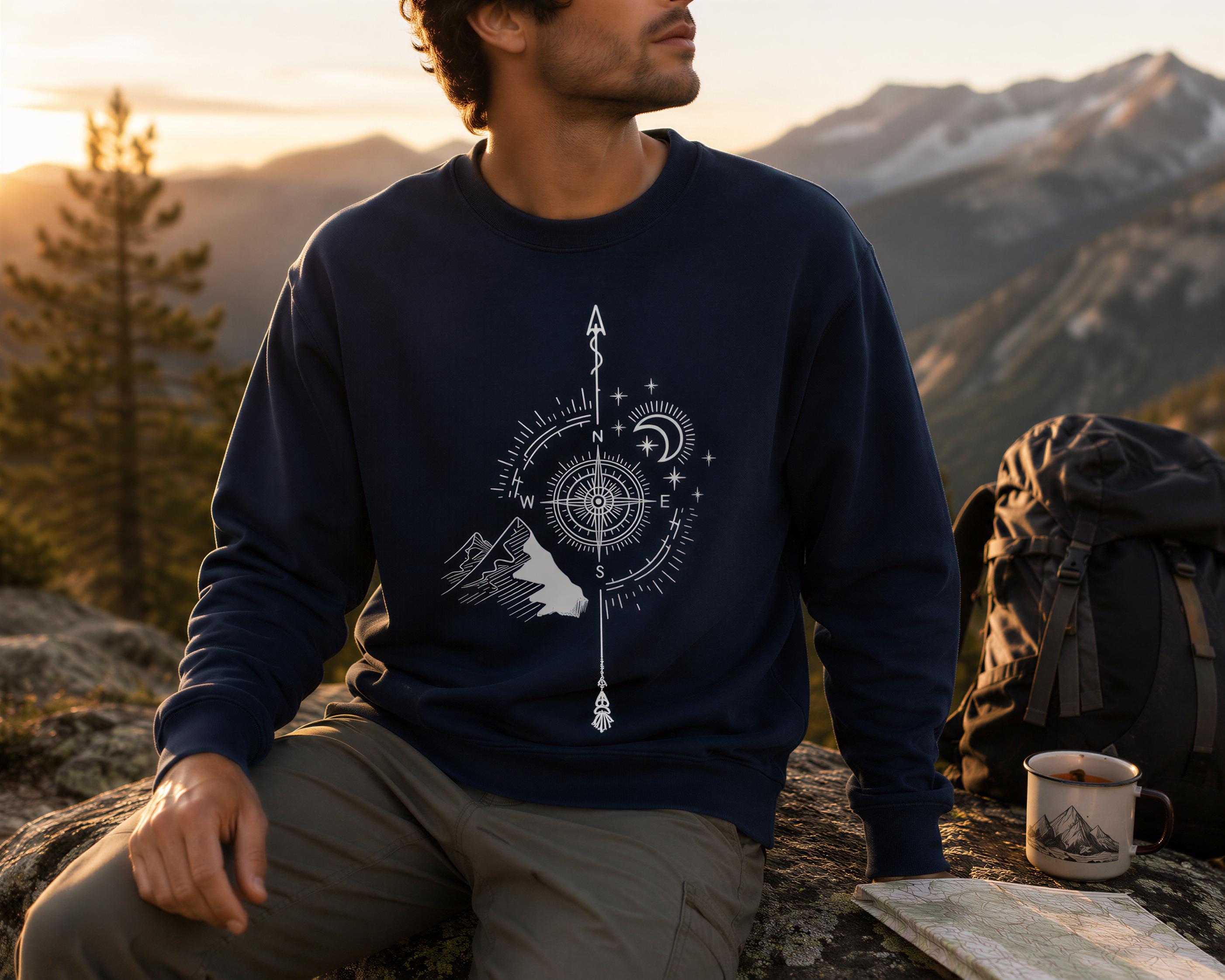 A man wearing a navy blue sweatshirt with a compass design sits on a rock, with a backpack and a cup of coffee nearby, against the backdrop of a mountain range at sunset.