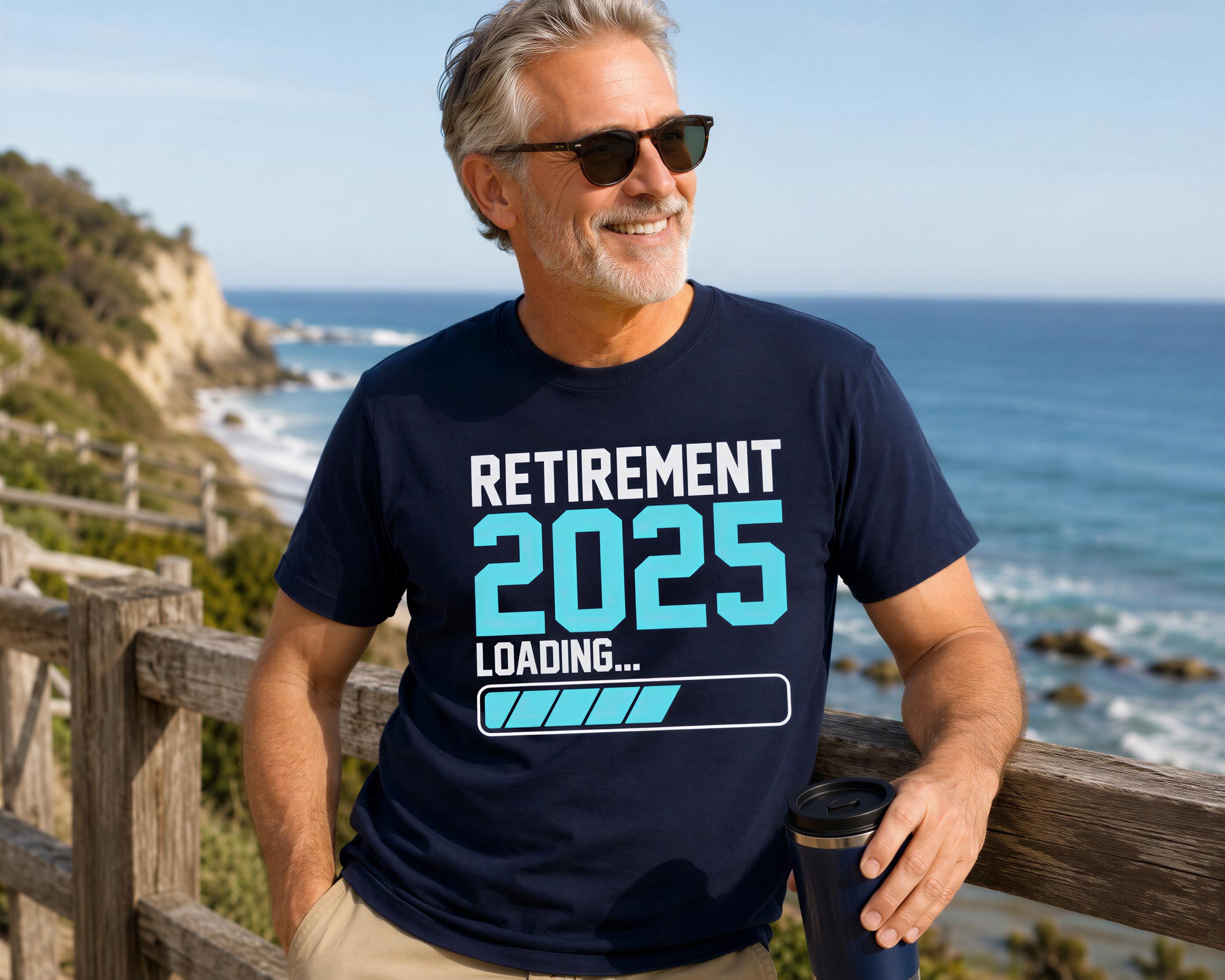 A man wearing a blue t-shirt with the text "Retirement 2025 Loading..." is standing on a wooden railing overlooking the ocean.