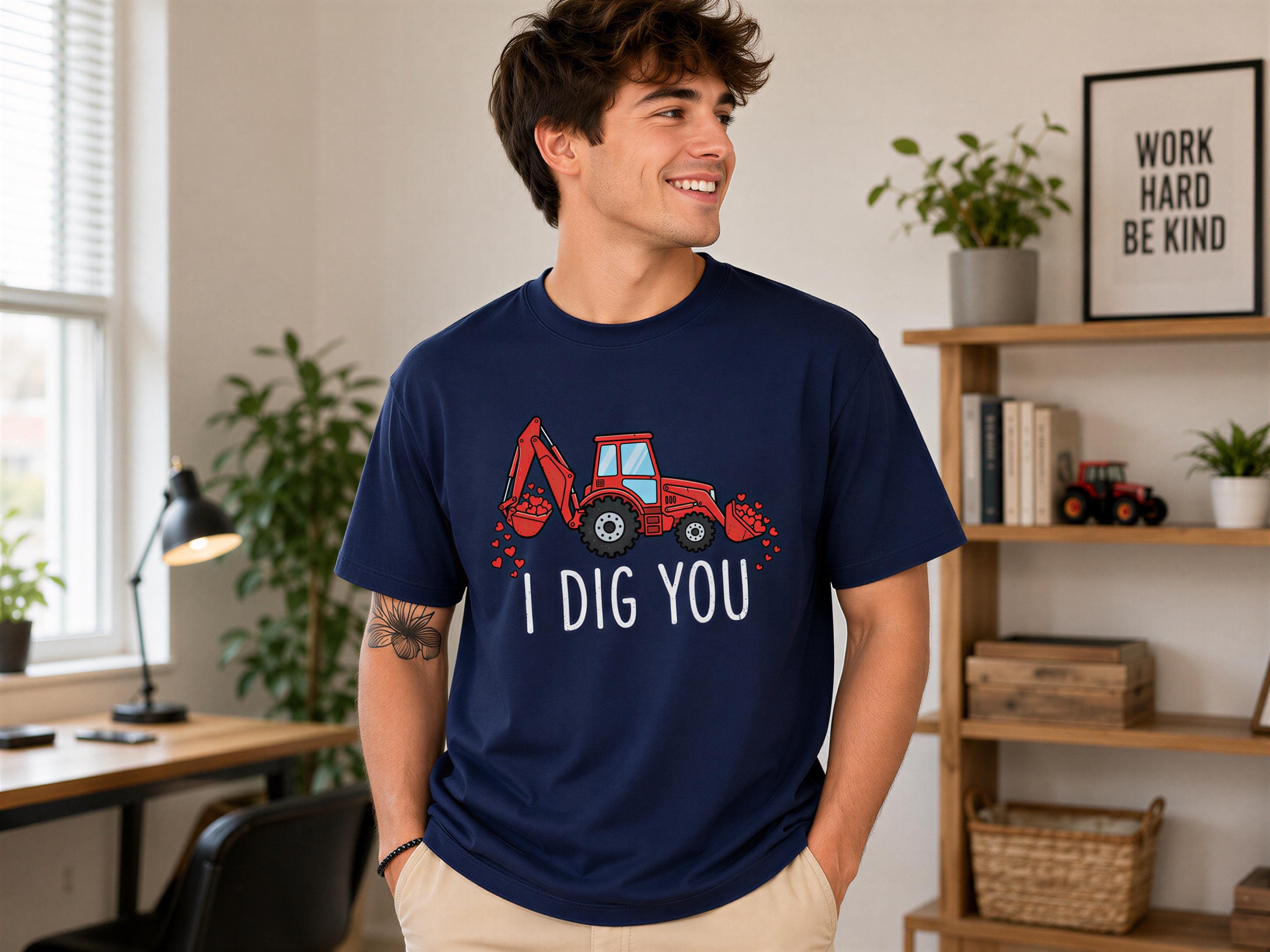 A young man wearing a navy blue t-shirt with a red tractor graphic and the text "I DIG YOU" stands in a room with a desk, chair, and potted plants.